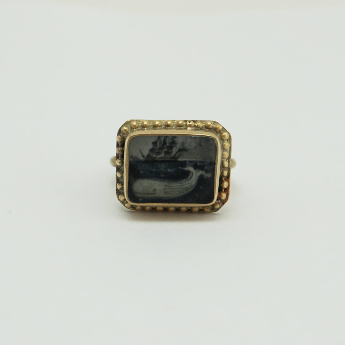 A gold rectangular ring with a hand painted whale, sea and ships. The outer part of the ring has circular bobbles around the edge.