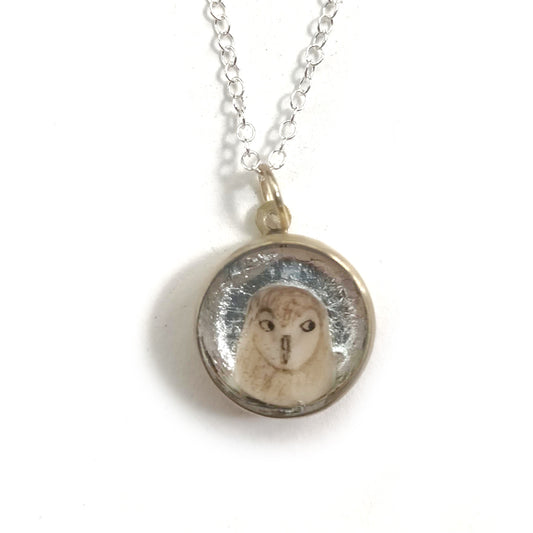A brass owl pendant with a hand painted insert on a silver background, attached to a sterling silver chain. The owl is white in colour and features some beige tones throughout. 