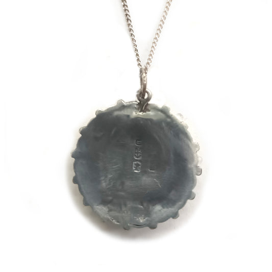 The back of a pendant, showing a shiny smooth silver surface attached to a silver fine chain. The back appears to feature some hallmarking symbols. 