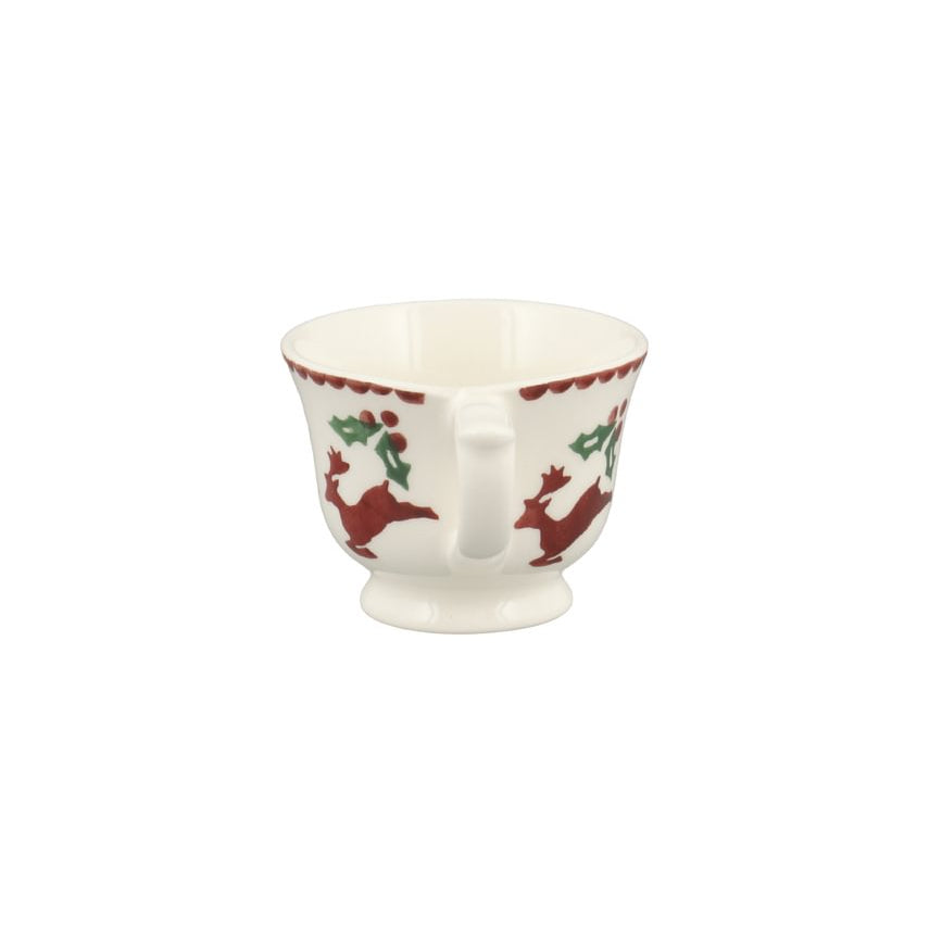 A small white ceramic cup with a handle and pedestal base, decorated with a red and green holiday pattern featuring deer and holly leaves.