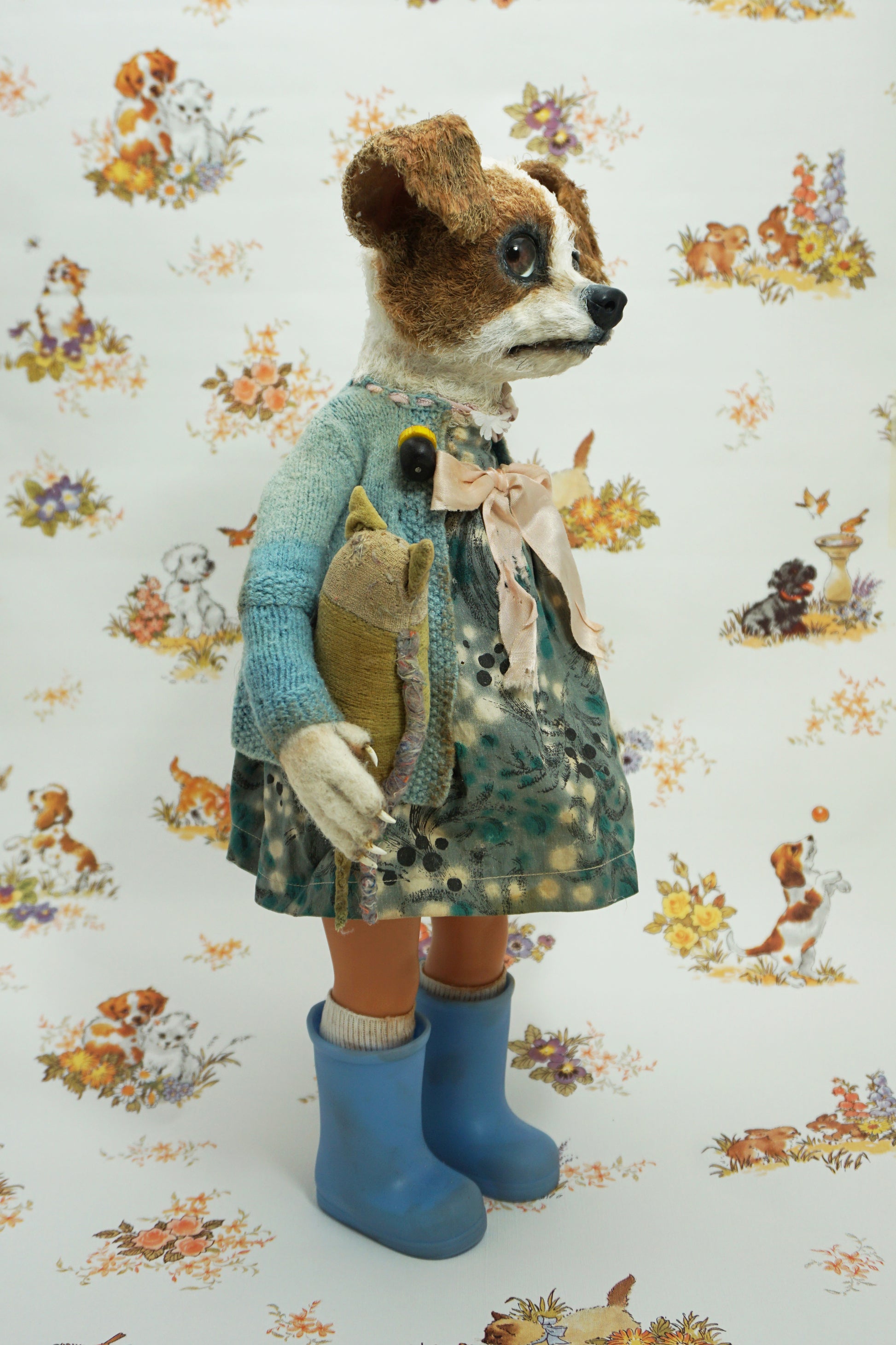 A whimsical doll resembling a dog stands upright, wearing a patterned dress, blue wellies, and a blue cardigan. It holds a small stuffed toy. The background features a playful animal print with various creatures.