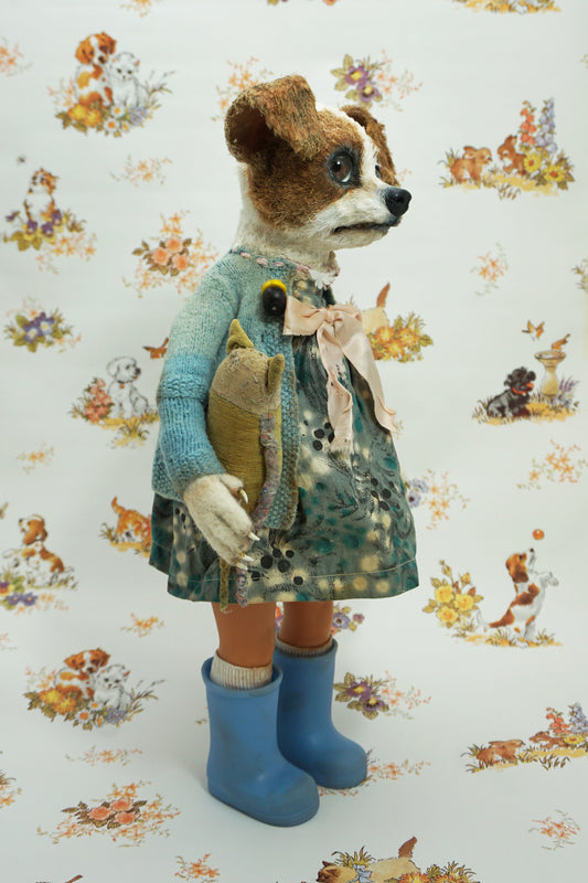 A whimsical doll resembling a dog stands upright, wearing a patterned dress, blue wellies, and a blue cardigan. It holds a small stuffed toy. The background features a playful animal print with various creatures.