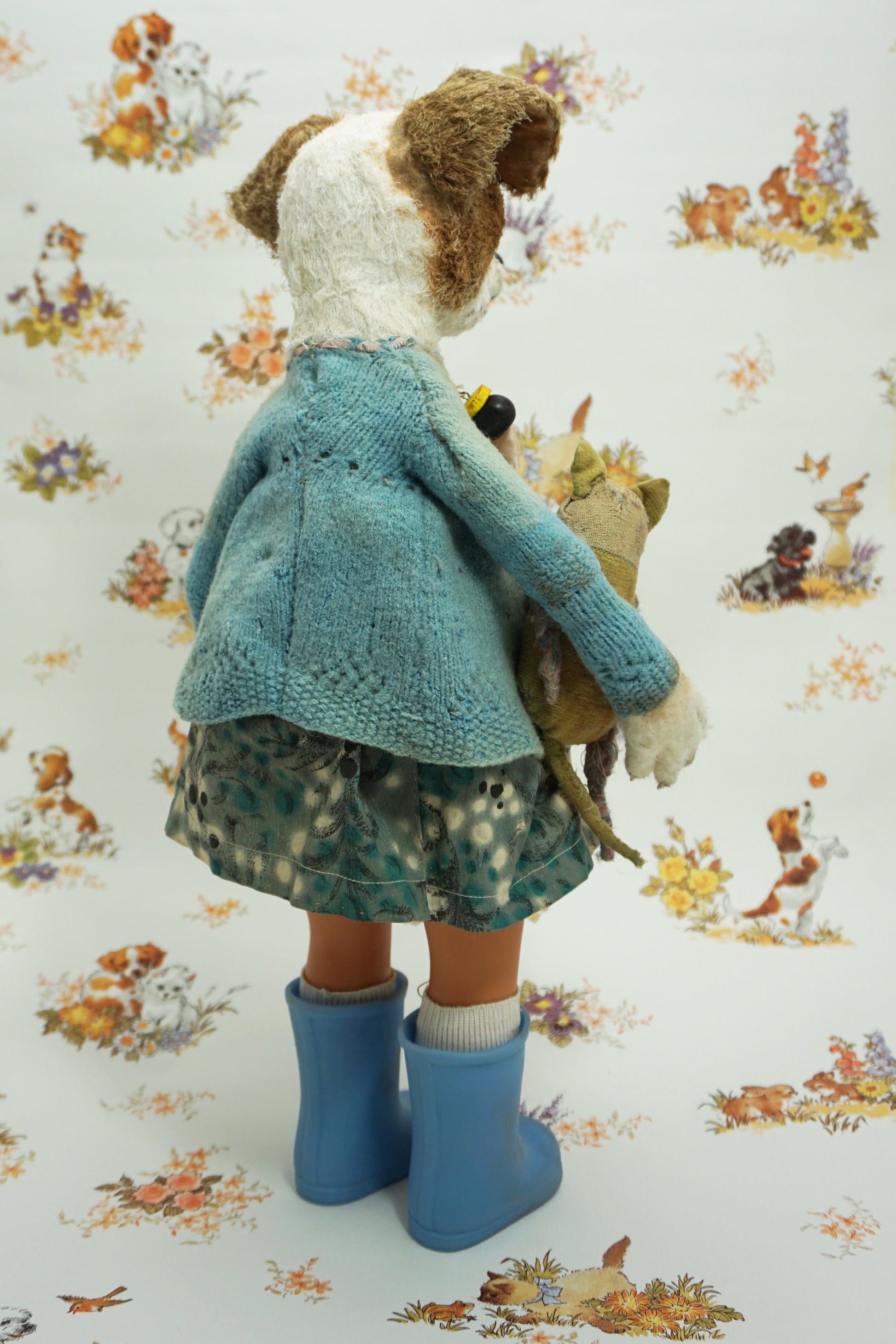 A whimsical doll resembling a dog stands upright, wearing a patterned dress, blue wellies, and a blue cardigan. It holds a small stuffed toy. The background features a playful animal print with various creatures.