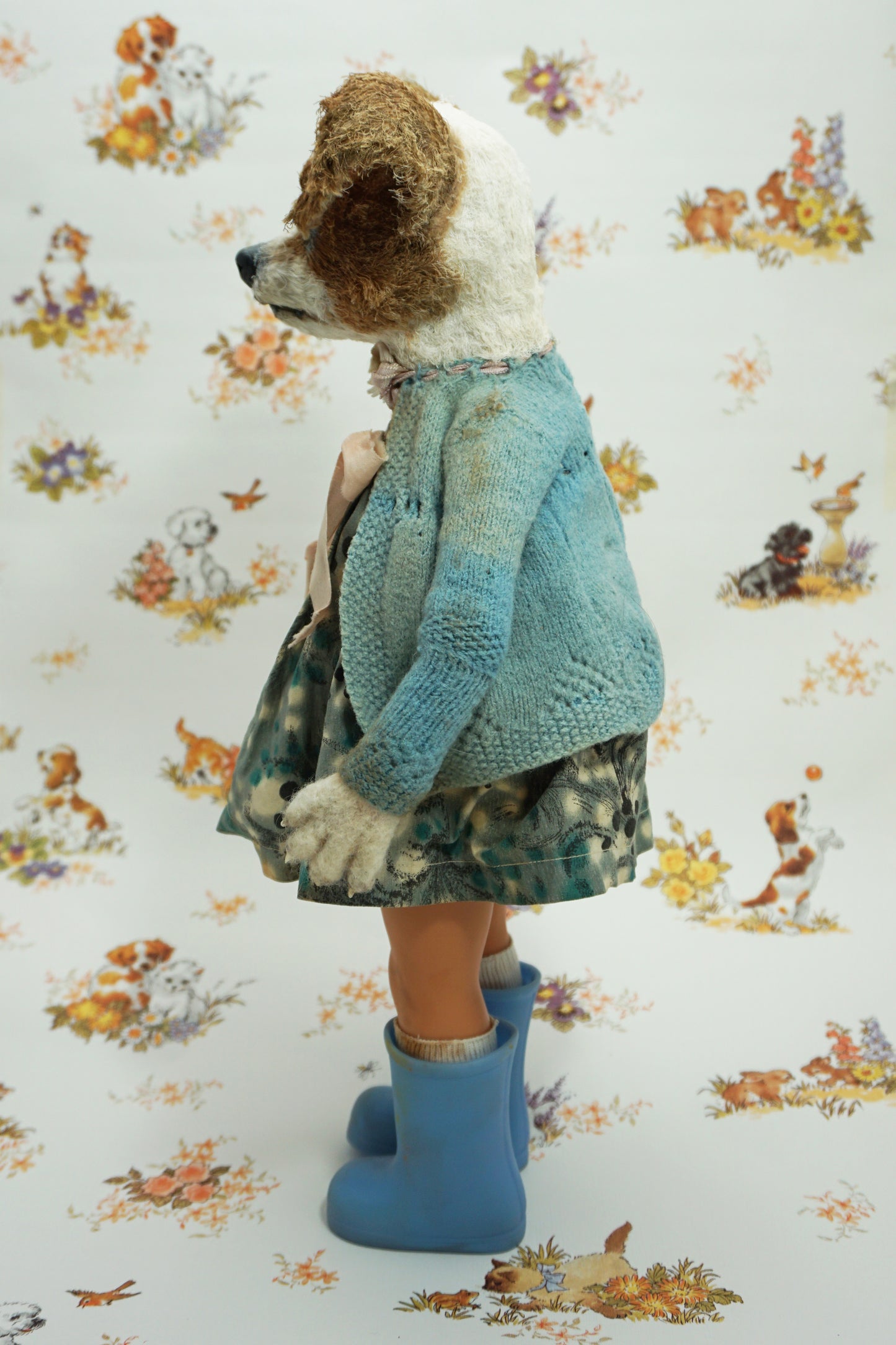 A whimsical doll resembling a dog stands upright, wearing a patterned dress, blue wellies, and a blue cardigan. It holds a small stuffed toy. The background features a playful animal print with various creatures.