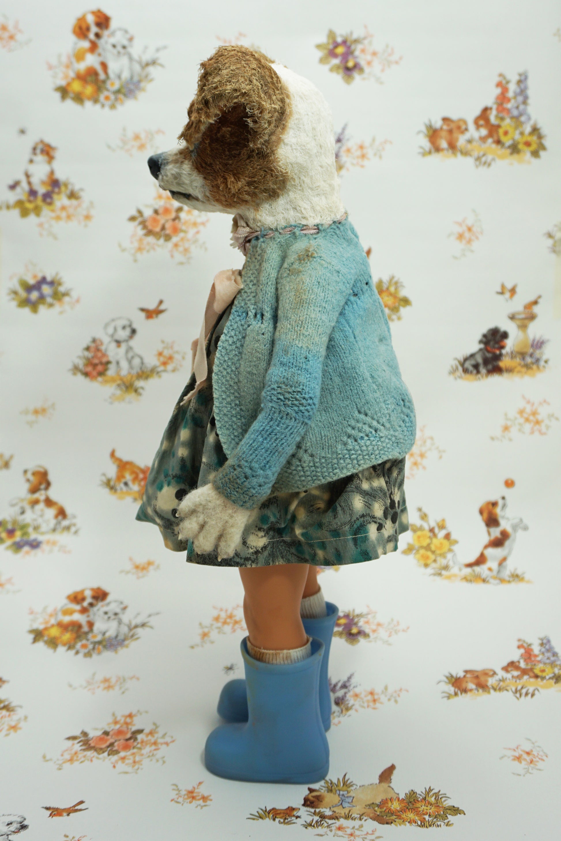 A whimsical doll resembling a dog stands upright, wearing a patterned dress, blue wellies, and a blue cardigan. It holds a small stuffed toy. The background features a playful animal print with various creatures.