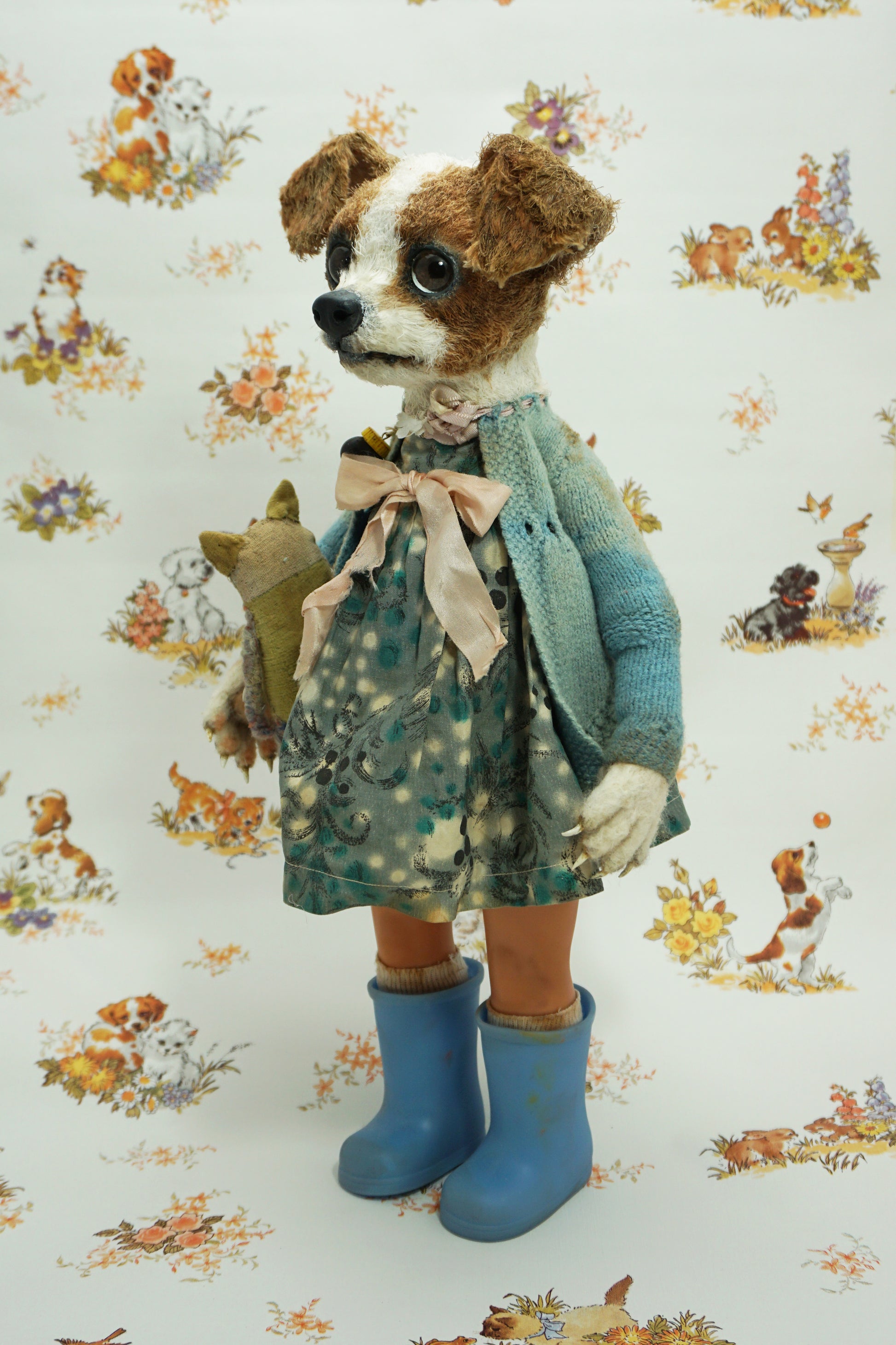 A whimsical doll resembling a dog stands upright, wearing a patterned dress, blue wellies, and a blue cardigan. It holds a small stuffed toy. The background features a playful animal print with various creatures.
