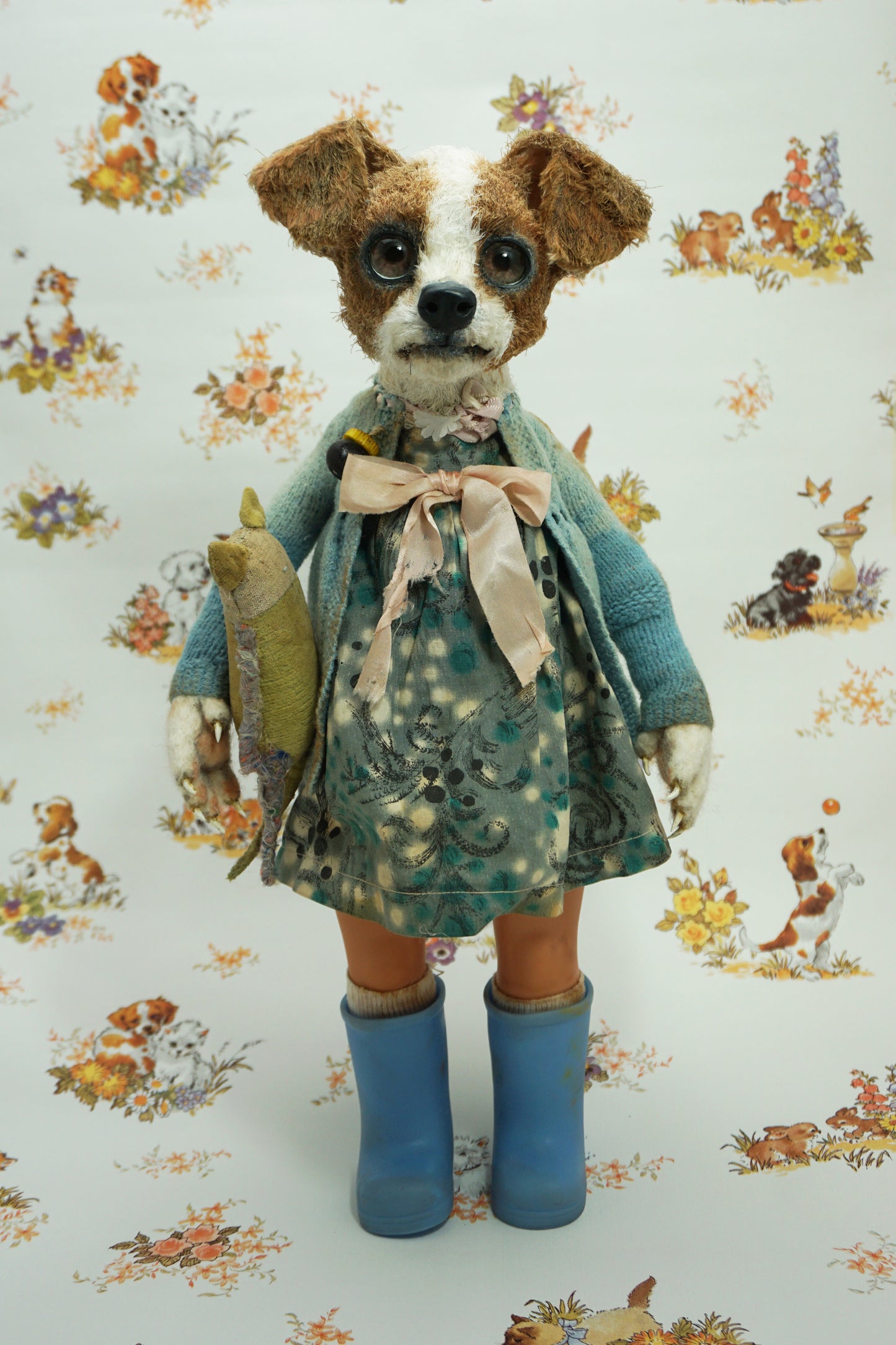 A whimsical doll resembling a dog stands upright, wearing a patterned dress, blue wellies, and a blue cardigan. It holds a small stuffed toy. The background features a playful animal print with various creatures.