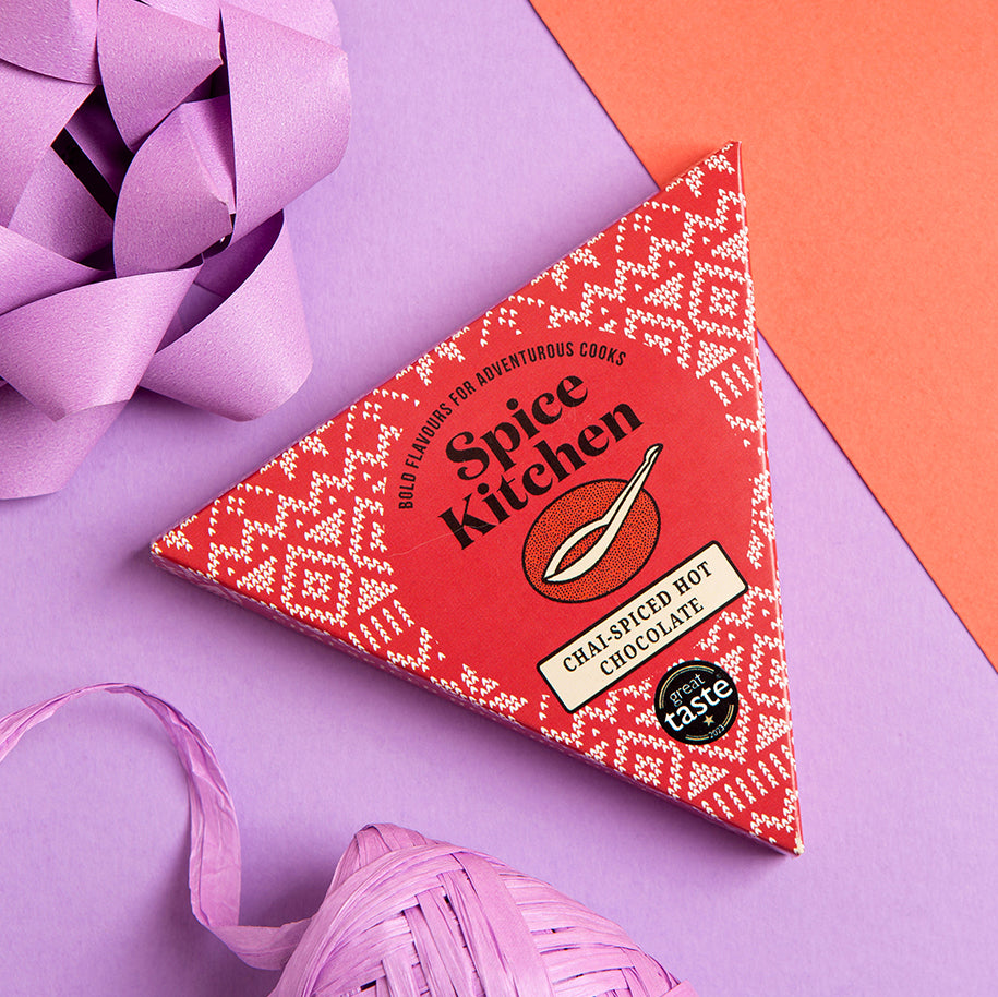 A triangular box of Spice Kitchen Chai Spiced Hot Chocolate, decorated with a red and white geometric pattern, styled on a purple and orange background with decorative paper and ribbon.