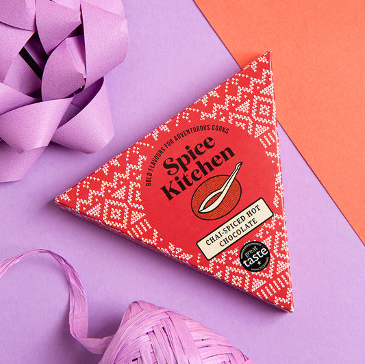 A triangular box of Spice Kitchen Chai Spiced Hot Chocolate, decorated with a red and white geometric pattern, styled on a purple and orange background with decorative paper and ribbon.