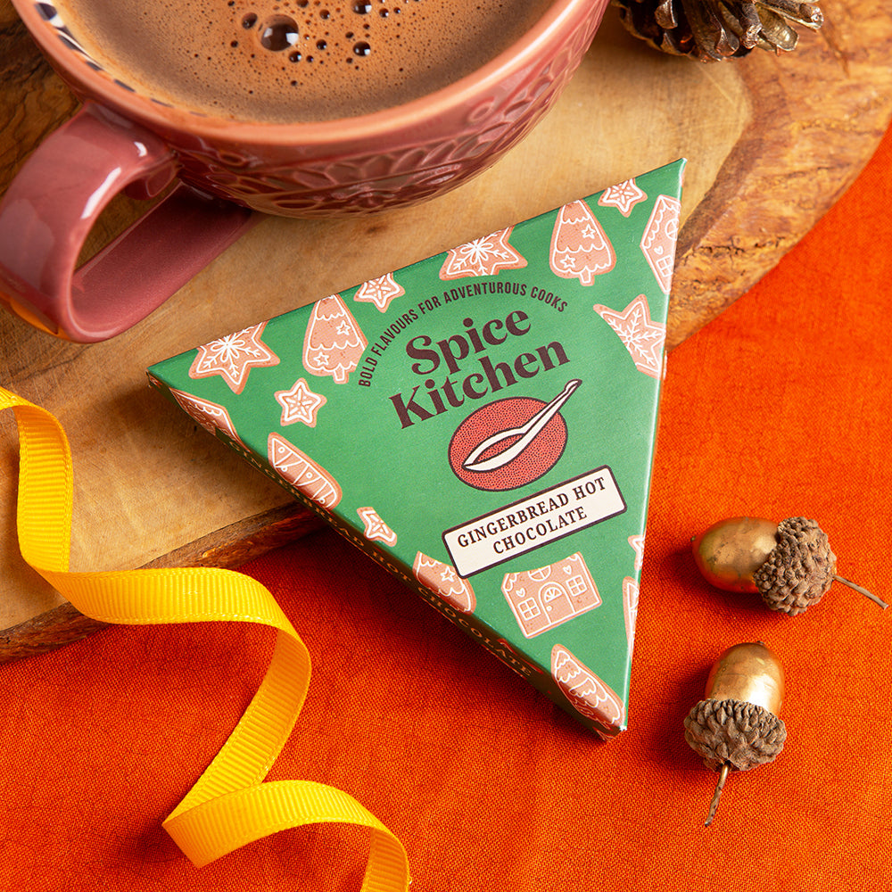 A triangular box of Spice Kitchen Gingerbread Hot Chocolate sits on a wooden board, styled with a pink mug of cocoa, a yellow ribbon, acorns, and an orange fabric background.