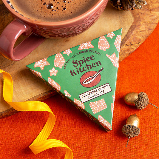 A triangular box of Spice Kitchen Gingerbread Hot Chocolate sits on a wooden board, styled with a pink mug of cocoa, a yellow ribbon, acorns, and an orange fabric background.