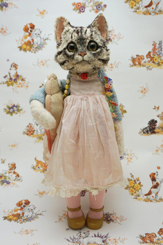 A plush grey tabby cat stands upright, wearing a pink dress, blue cardigan, and holding a fish soft toy. It has large eyes and a red bead for a collar. The background features a floral and animal pattern with small dogs and flowers.