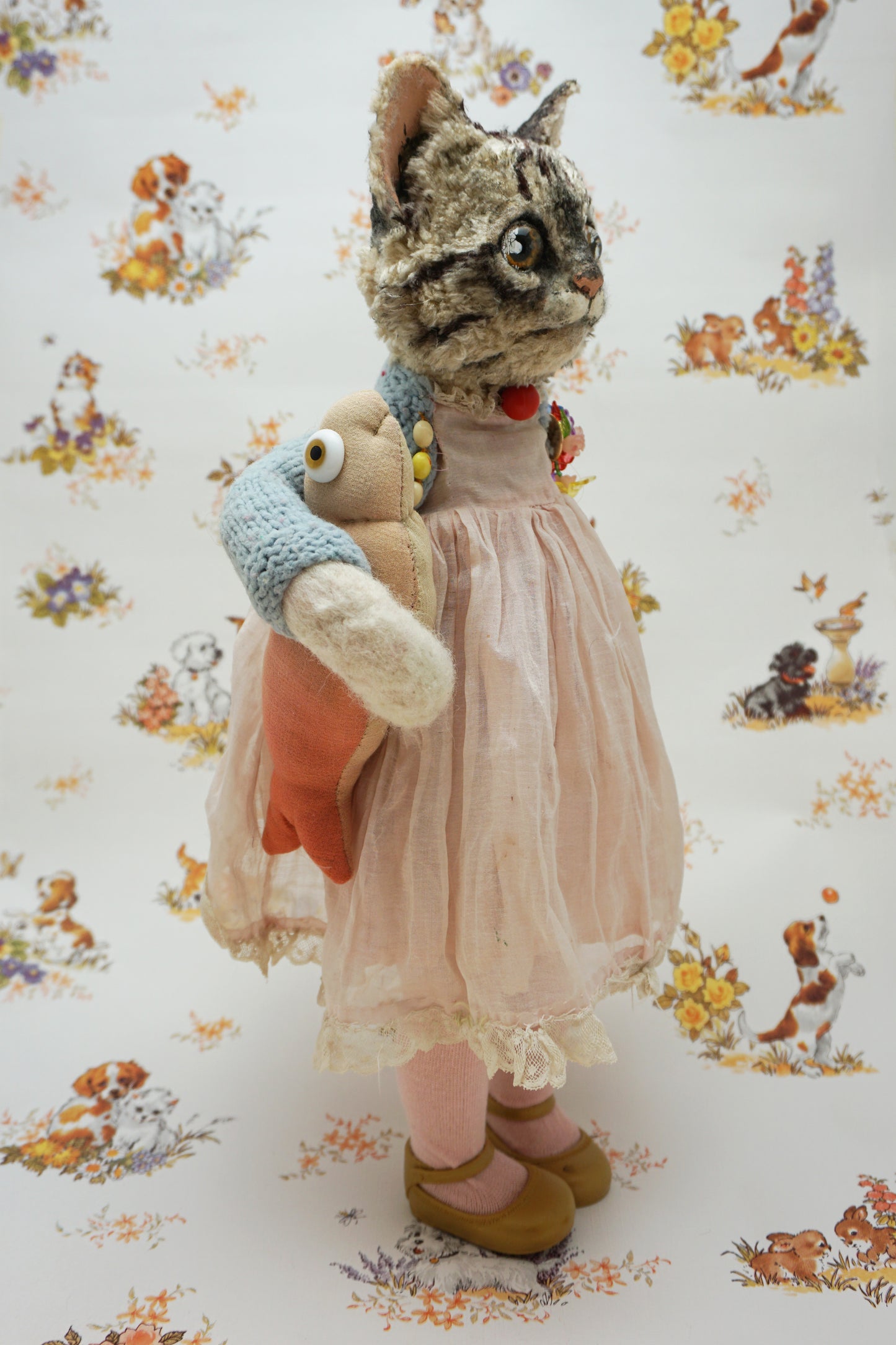 A plush grey tabby cat stands upright, wearing a pink dress, blue cardigan, and holding a fish soft toy. It has large eyes and a red bead for a collar. The background features a floral and animal pattern with small dogs and flowers.