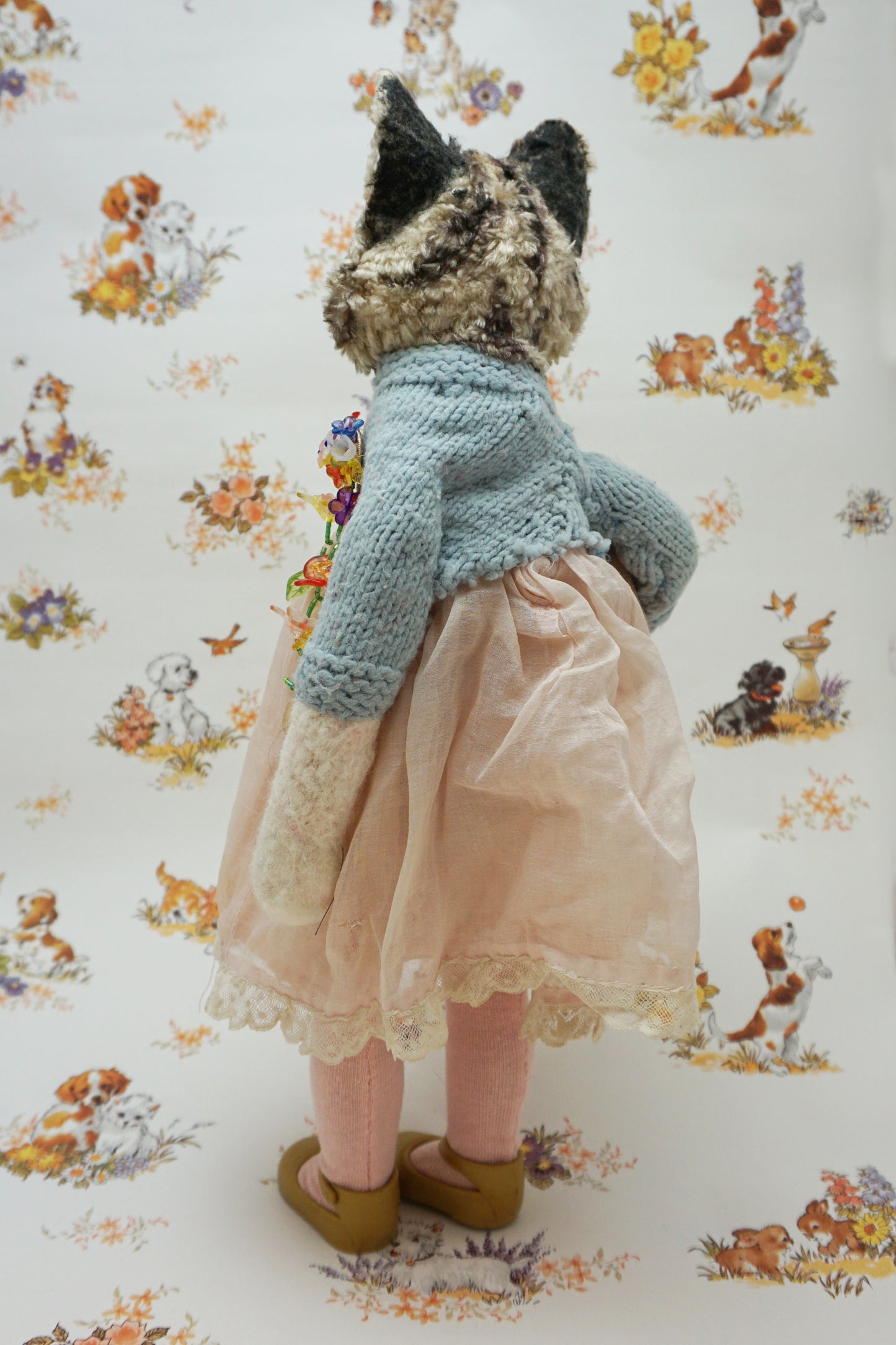 A plush grey tabby cat stands upright, wearing a pink dress, blue cardigan, and holding a fish soft toy. It has large eyes and a red bead for a collar. The background features a floral and animal pattern with small dogs and flowers.