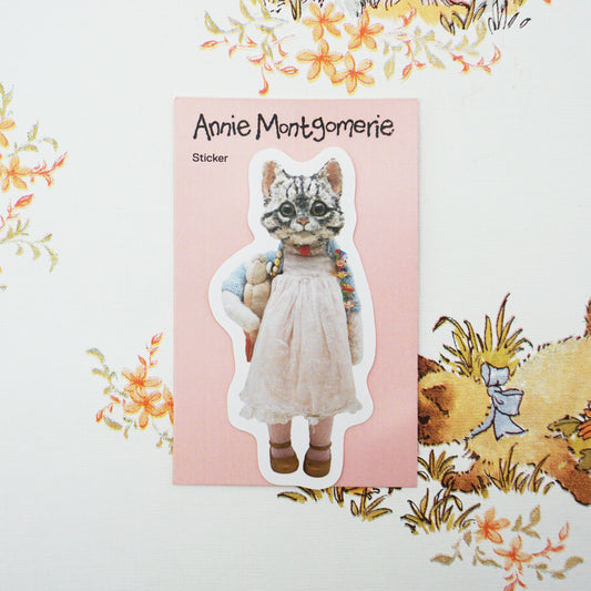 A die-cut sticker featuring a cat shown on its pink backing card labelled 'Annie Montgomerie Sticker'. The card is placed on a vintage floral-patterned surface.