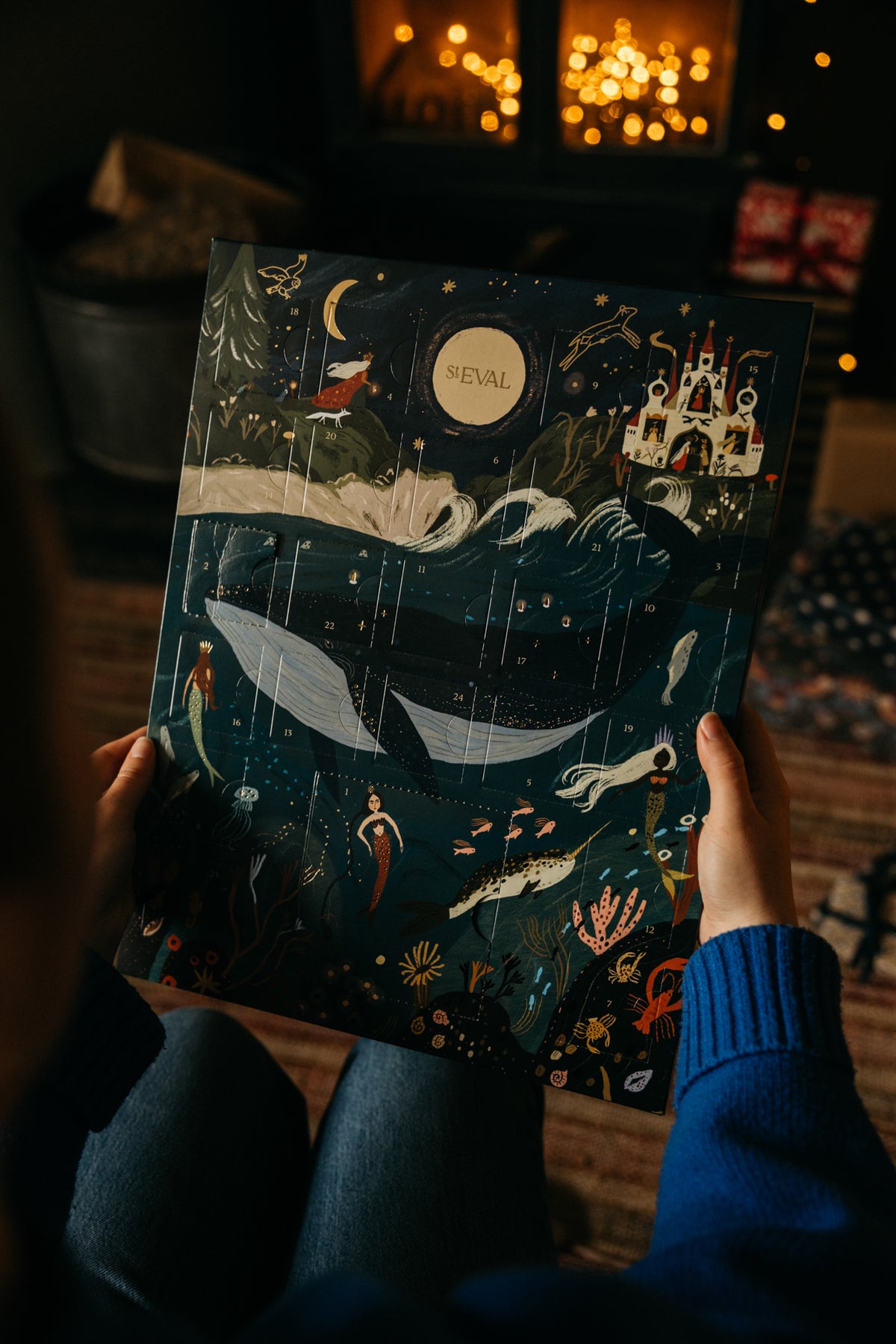 A person holds a dark illustrated advent calendar featuring whimsical sea creatures, a castle, and a moon with the word 'St Eval' visible. The scene is photographed in a cosy indoor setting with warm blurred lights in the background.