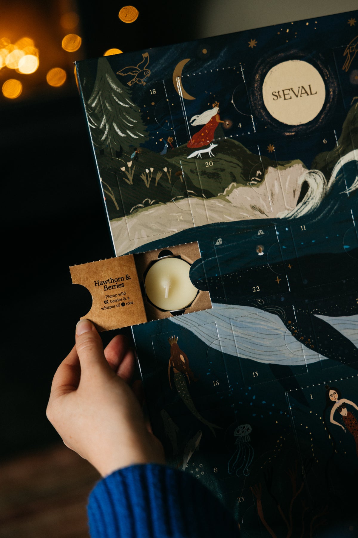 A hand holds a ST EVAL advent calendar with a winter fantasy illustration, showing a small candle peeking out from a cardboard flap. The scene is lit by warm, blurred bokeh lights in the background.