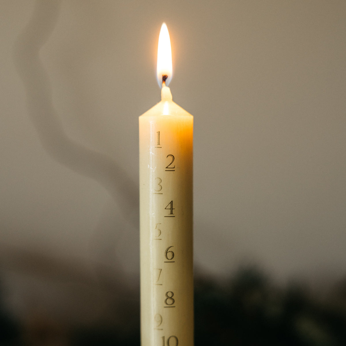 A single lit cream-colored taper candle with black printed numbers from 1 to 24 running vertically, shown against a softly blurred dark background.