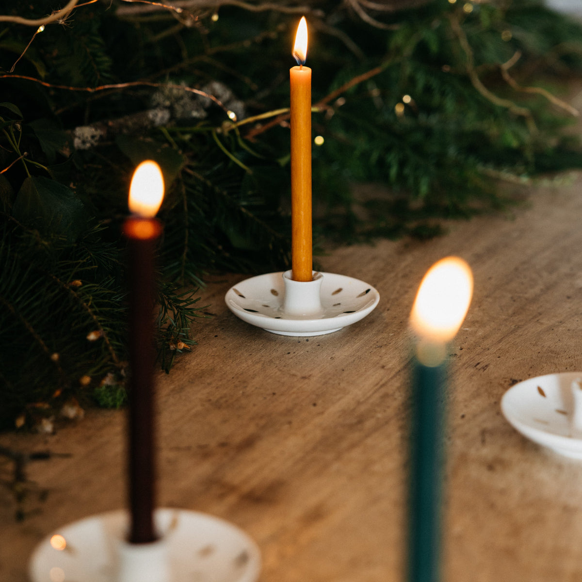 Three lit taper candles in white holders on a wooden surface, with greenery and warm bokeh lights in the background.