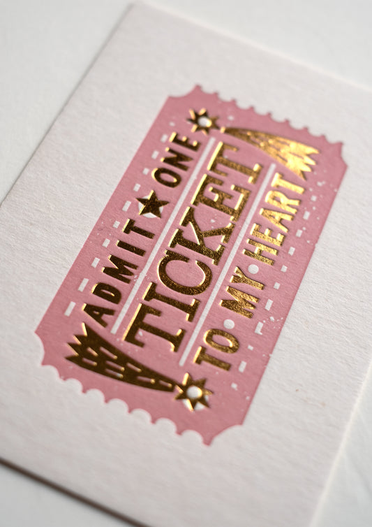 A pink ticket design featuring the text "Admit One Ticket to My Heart" in bold, decorative lettering. The ticket has a vintage style with stars and decorative elements, set against a textured white background.