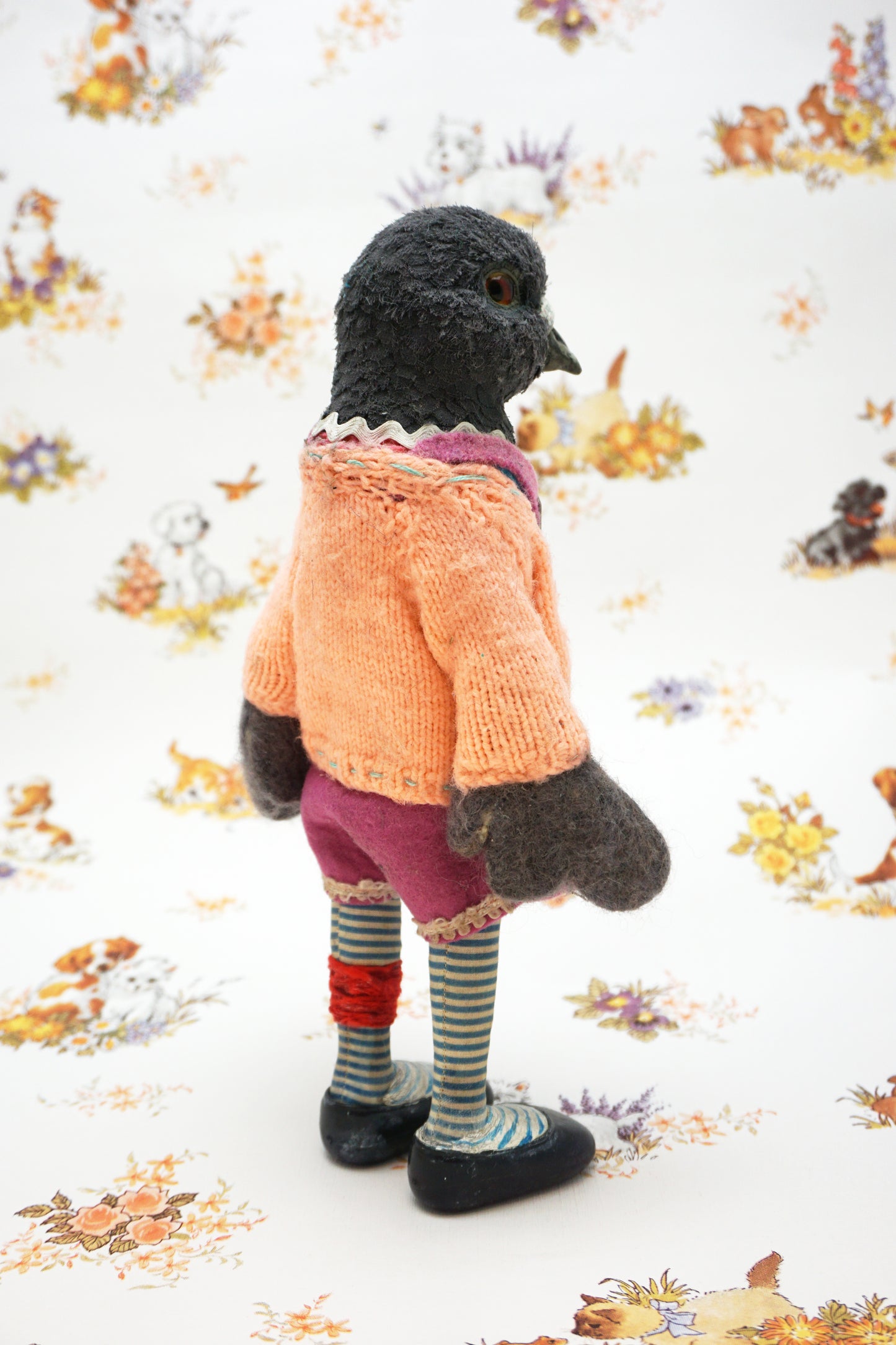 A plush pigeon dressed in a pink dress, striped leggings, and a peach cardigan with colourful buttons stands against a whimsical background featuring small animal illustrations and floral patterns.