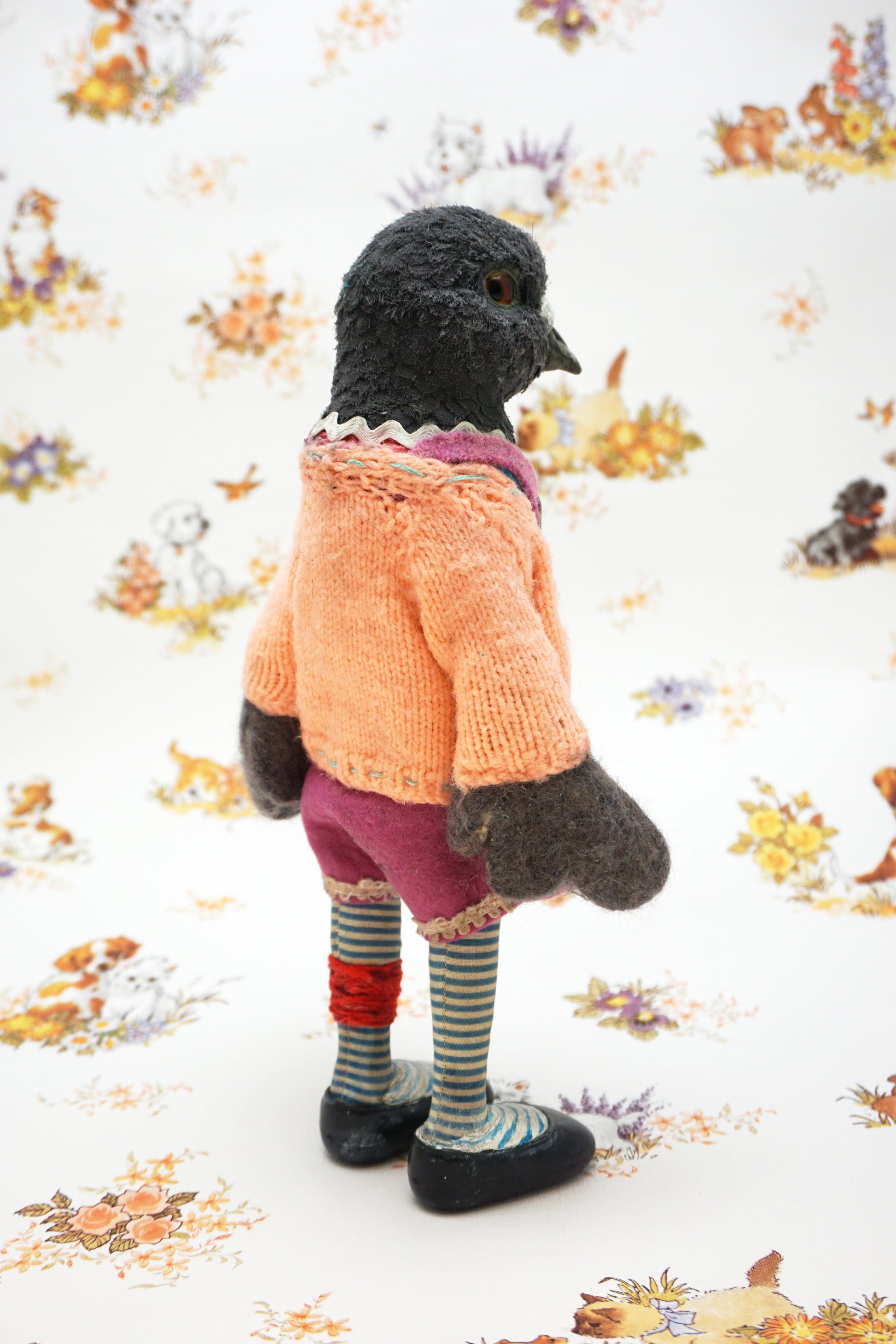 A plush pigeon dressed in a pink dress, striped leggings, and a peach cardigan with colourful buttons stands against a whimsical background featuring small animal illustrations and floral patterns.