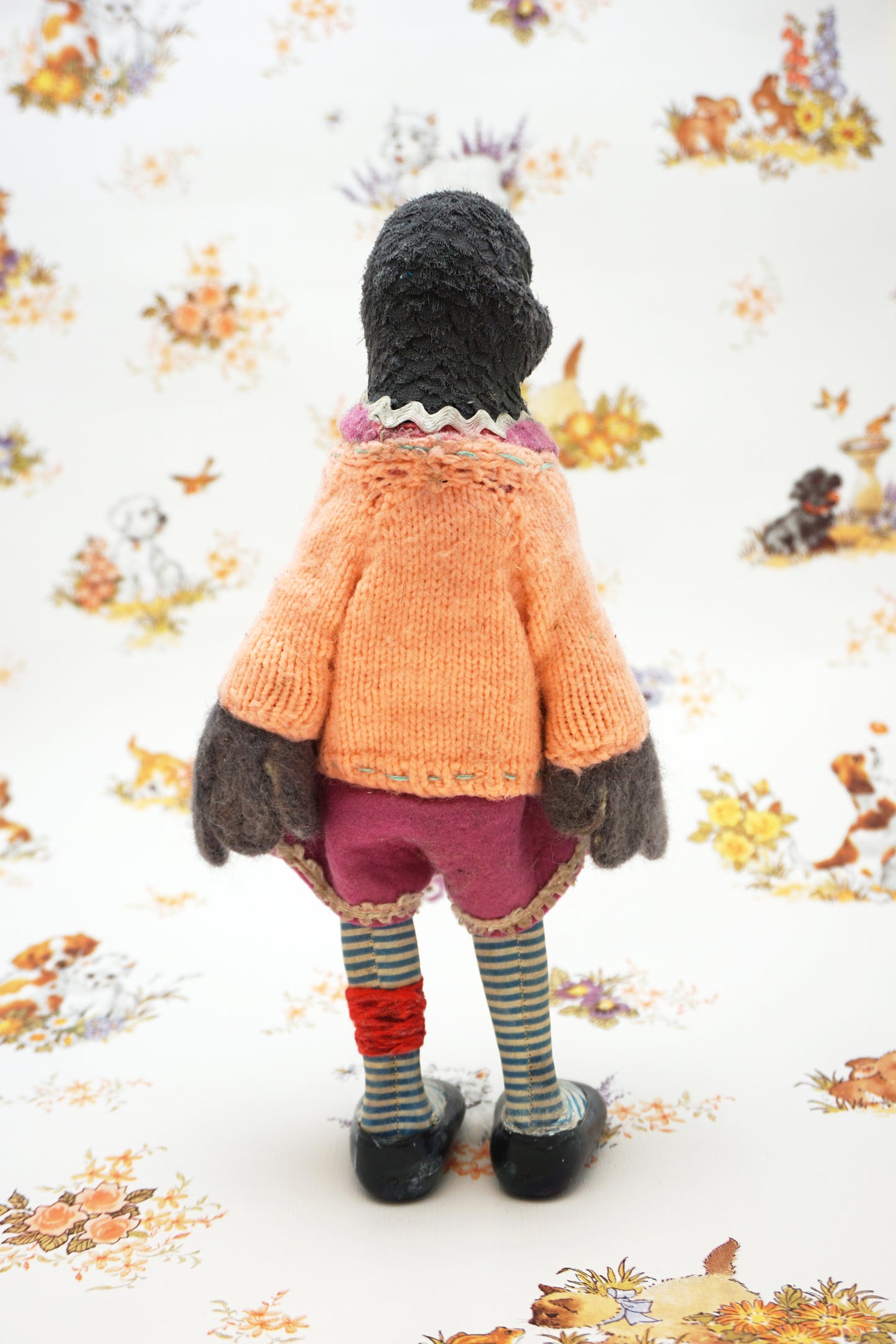 A plush pigeon dressed in a pink dress, striped leggings, and a peach cardigan with colourful buttons stands against a whimsical background featuring small animal illustrations and floral patterns.