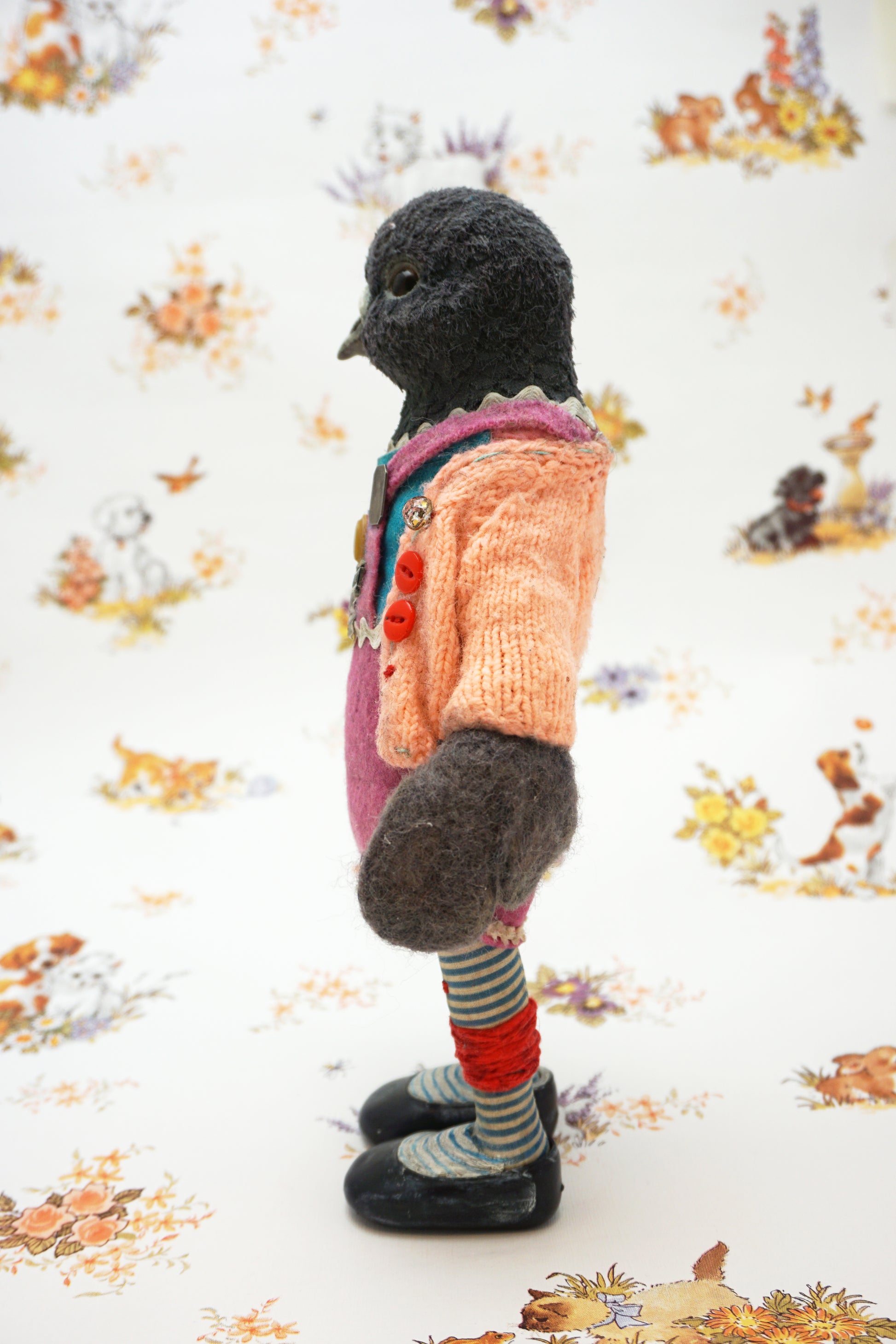 A plush pigeon dressed in a pink dress, striped leggings, and a peach cardigan with colourful buttons stands against a whimsical background featuring small animal illustrations and floral patterns.