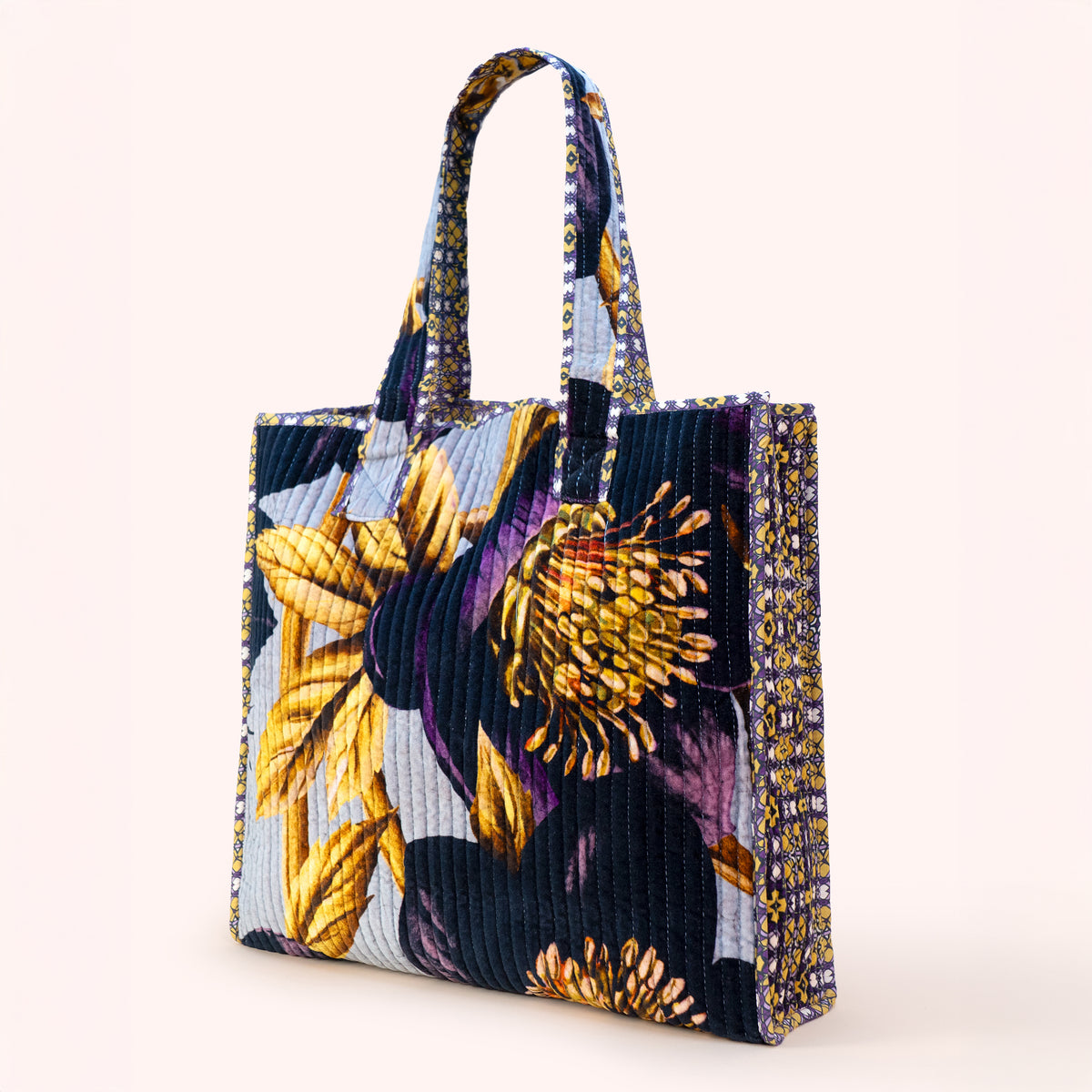 A structured tote bag featuring a textured velvet fabric with a floral print in yellow, purple, and blue, set against a solid pale pink background.