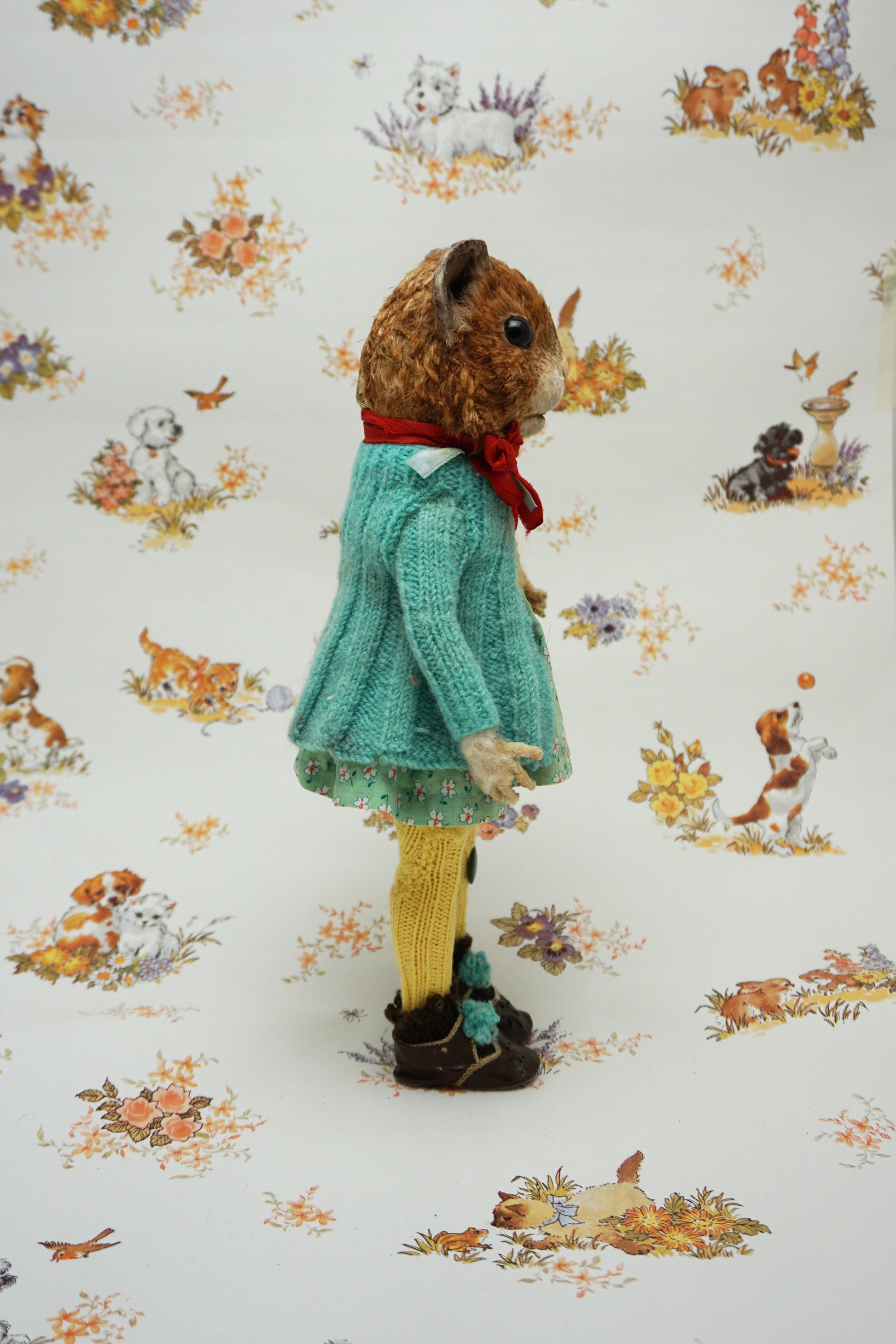 A plush guinea pig dressed in a teal cardigan, floral dress, and yellow tights holds a small stuffed animal. The background features whimsical illustrations of animals and nature scenes.