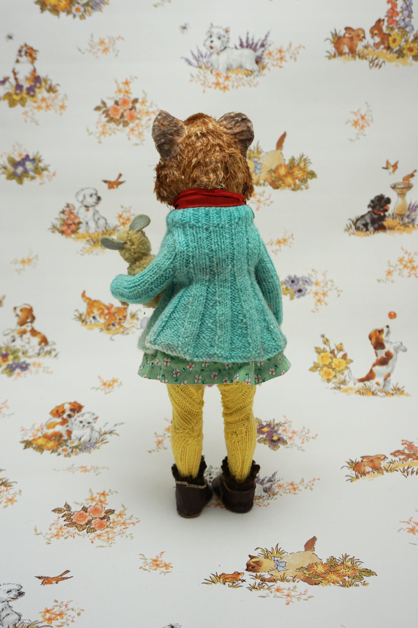 A plush guinea pig dressed in a teal cardigan, floral dress, and yellow tights holds a small stuffed animal. The background features whimsical illustrations of animals and nature scenes.