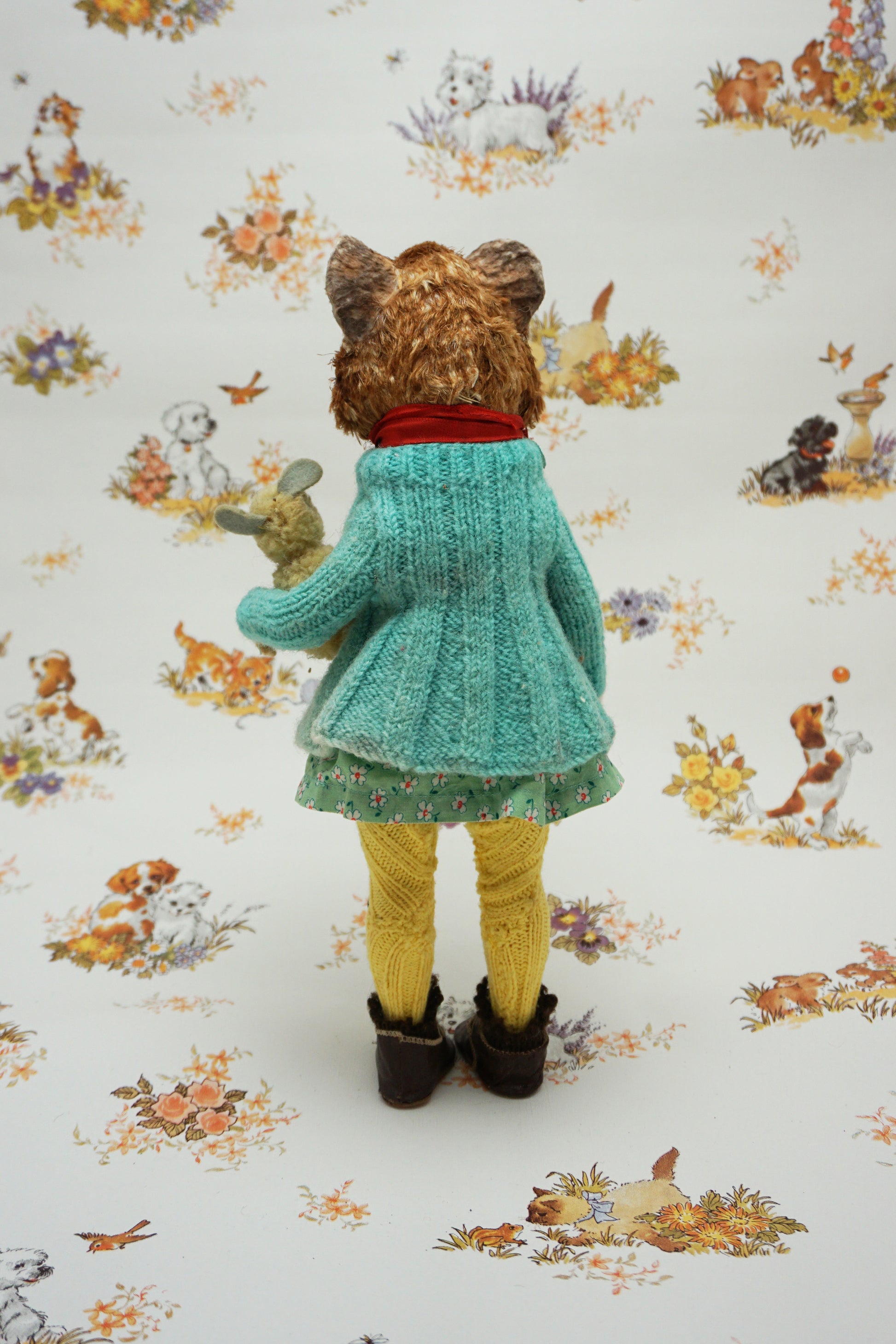 A plush guinea pig dressed in a teal cardigan, floral dress, and yellow tights holds a small stuffed animal. The background features whimsical illustrations of animals and nature scenes.