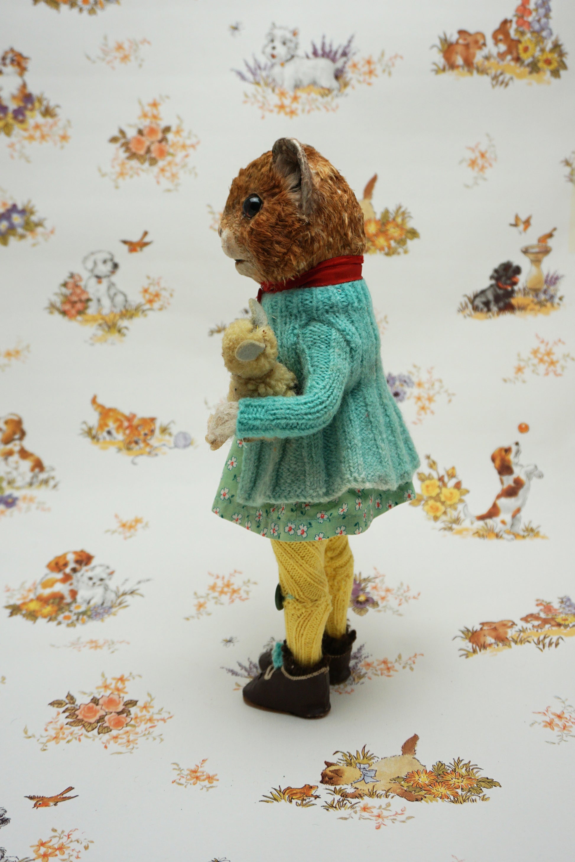 A plush guinea pig dressed in a teal cardigan, floral dress, and yellow tights holds a small stuffed animal. The background features whimsical illustrations of animals and nature scenes.
