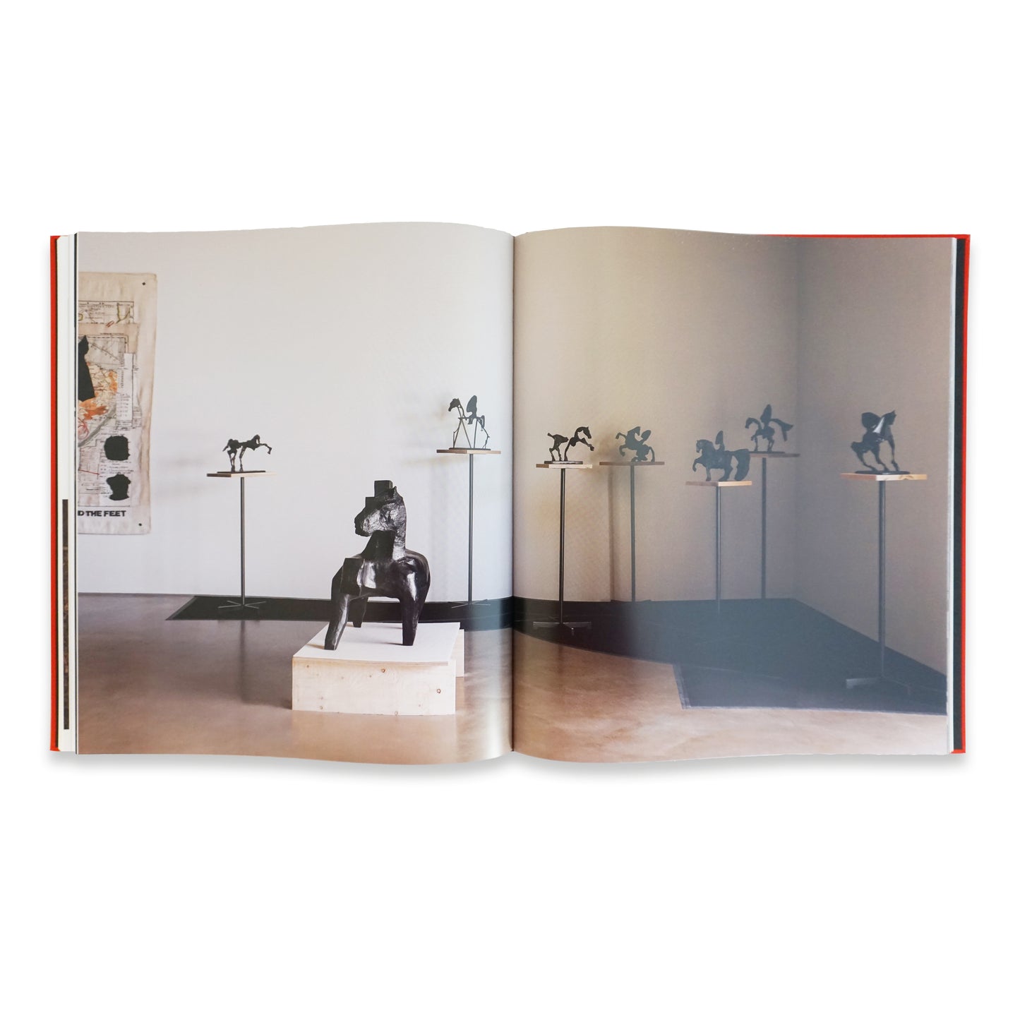 A spacious gallery featuring an open book with sculptures displayed on pedestals. The sculptures depict various figures, with a prominent piece on a block in the foreground. The walls are minimalistic, enhancing the artwork's focus.