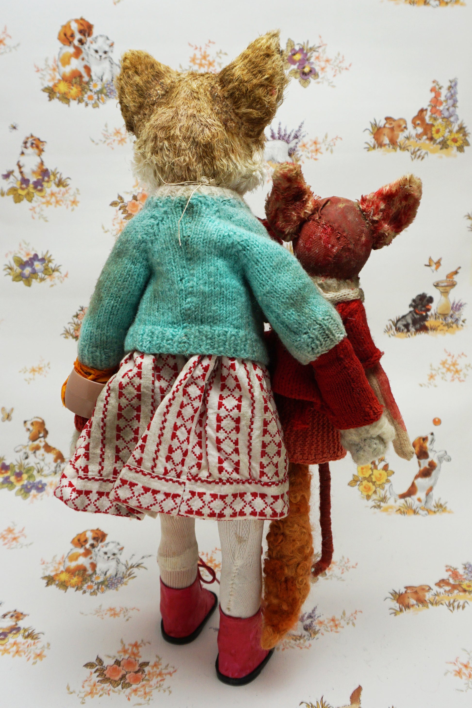 A plush fox toy dressed in a patterned red and white dress, teal cardigan, and red boots holds a smaller red plush creature. The background features a whimsical animal print with various small animals.