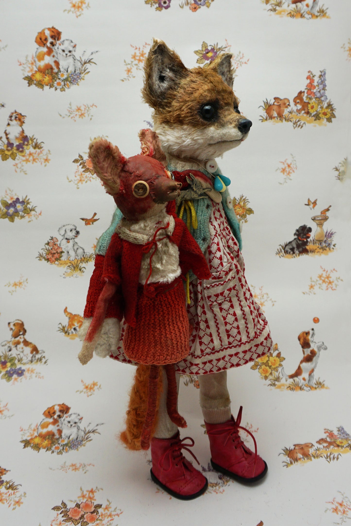 A plush fox toy dressed in a patterned red and white dress, teal cardigan, and red boots holds a smaller red plush creature. The background features a whimsical animal print with various small animals.