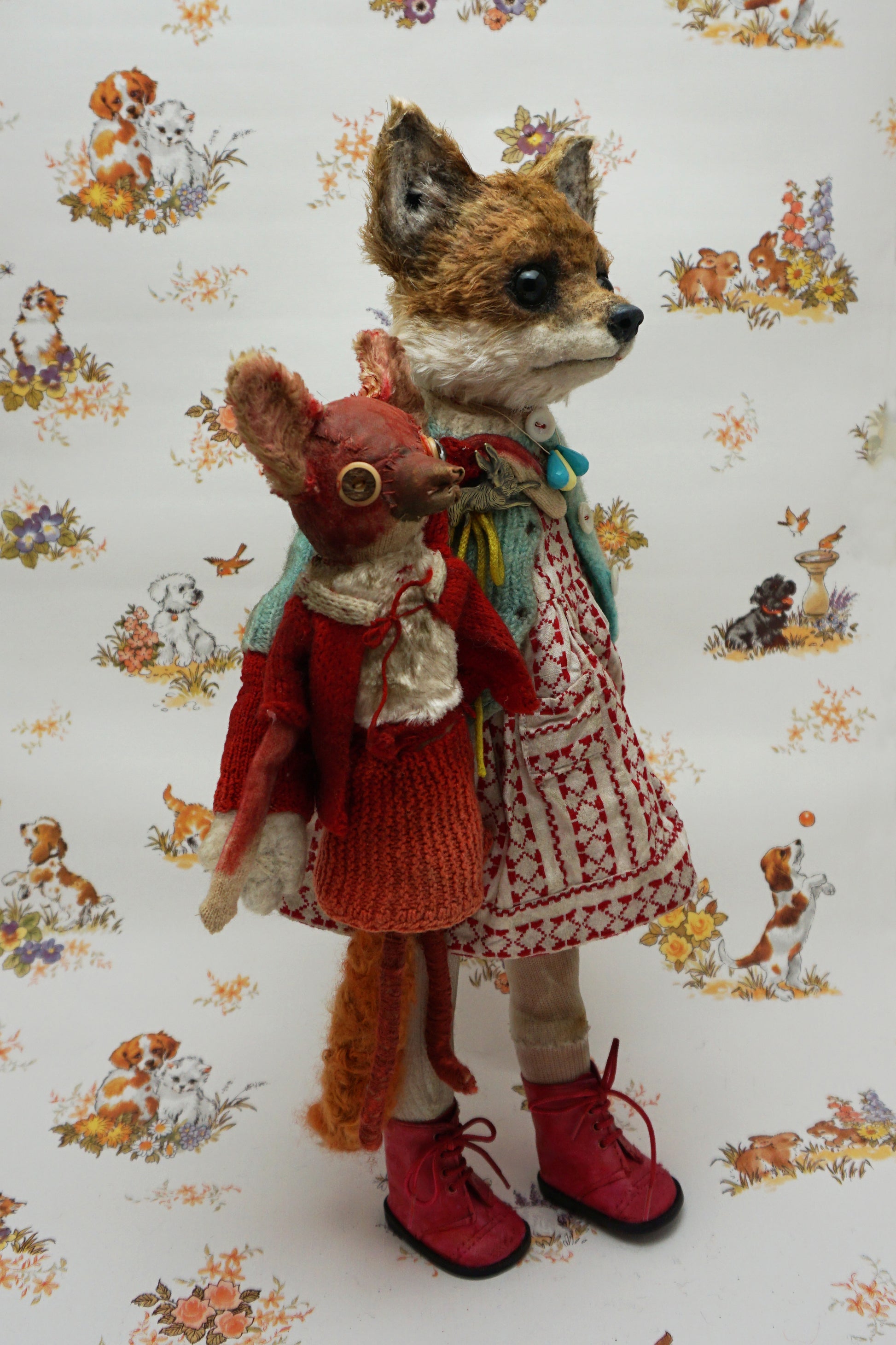 A plush fox toy dressed in a patterned red and white dress, teal cardigan, and red boots holds a smaller red plush creature. The background features a whimsical animal print with various small animals.