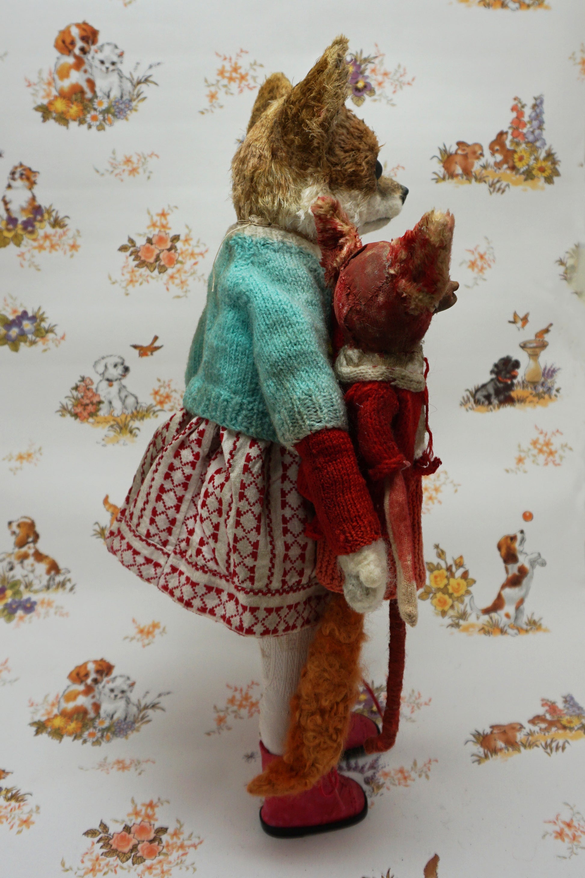 A plush fox toy dressed in a patterned red and white dress, teal cardigan, and red boots holds a smaller red plush creature. The background features a whimsical animal print with various small animals.