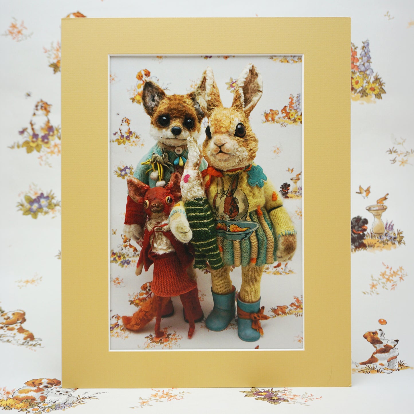 A mini print of two plush animals: a fox and a rabbit. They are dressed in colourful, knitted clothing. The background features a whimsical pattern with similar animal illustrations. The print has a yellow mount.