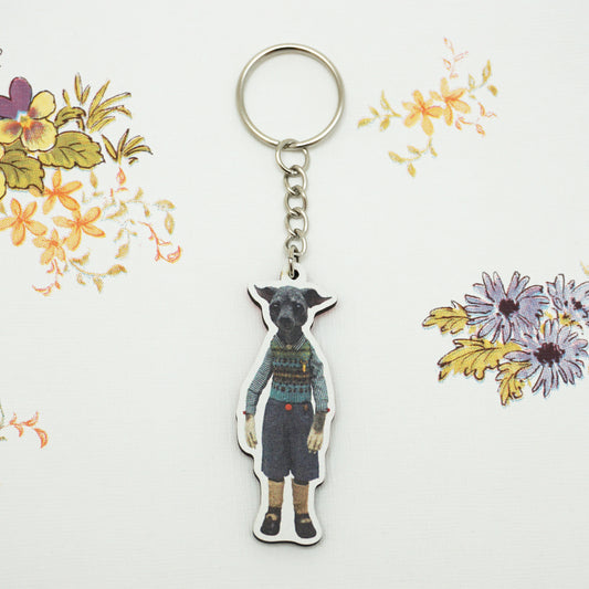 A keyring featuring a cut-out image of a dog dressed in vintage-style human clothing, including a knitted vest and shorts. The background is a floral-patterned fabric with various colourful flowers.