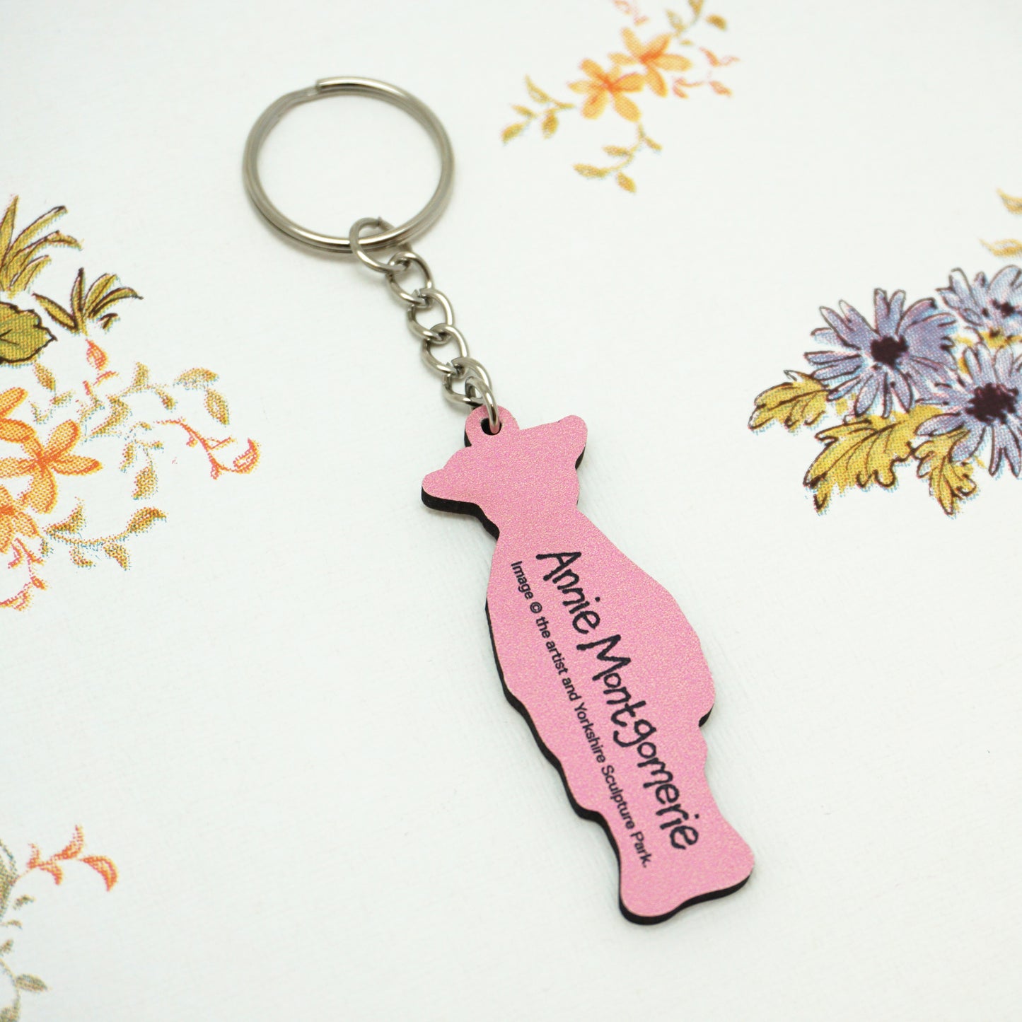The back of the Wilton keyring is pink with the text "Annie Montgomerie" printed on it. The keyring is placed on a white surface with a floral pattern featuring orange and purple flowers.