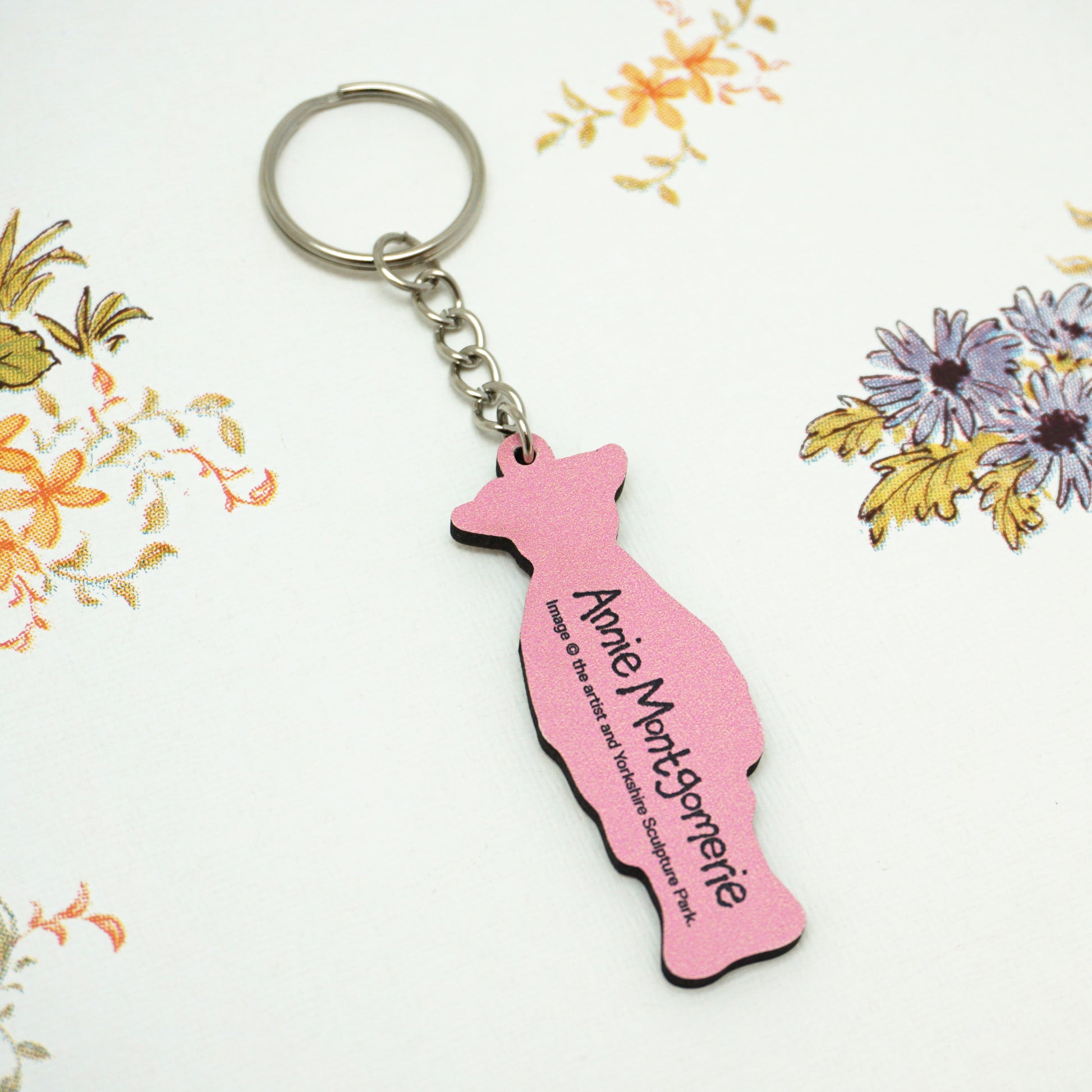 The back of the Wilton keyring is pink with the text "Annie Montgomerie" printed on it. The keyring is placed on a white surface with a floral pattern featuring orange and purple flowers.