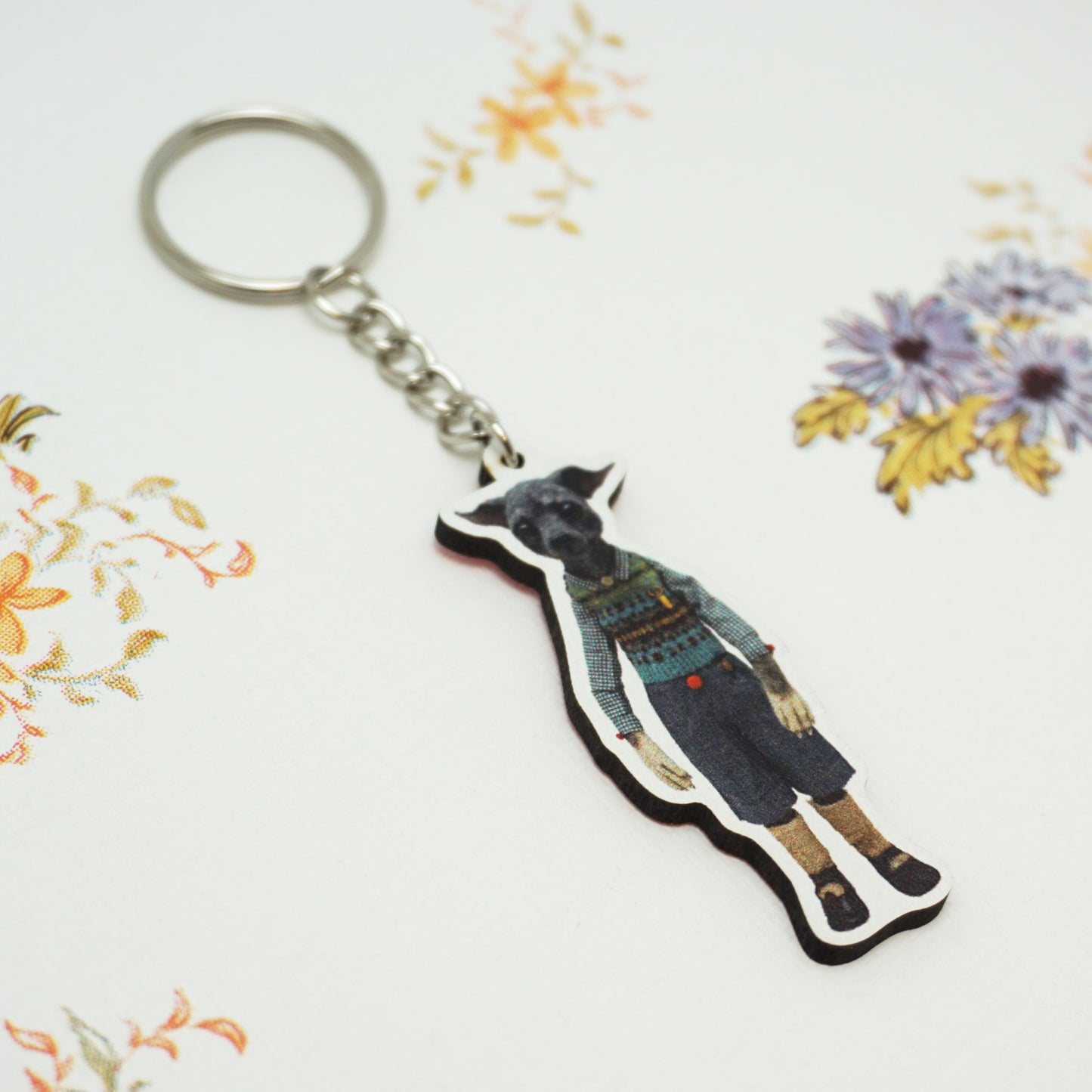 A keyring featuring a cut-out figure of a dog in vintage clothing. The background is a white surface with floral patterns in orange, purple, and yellow.