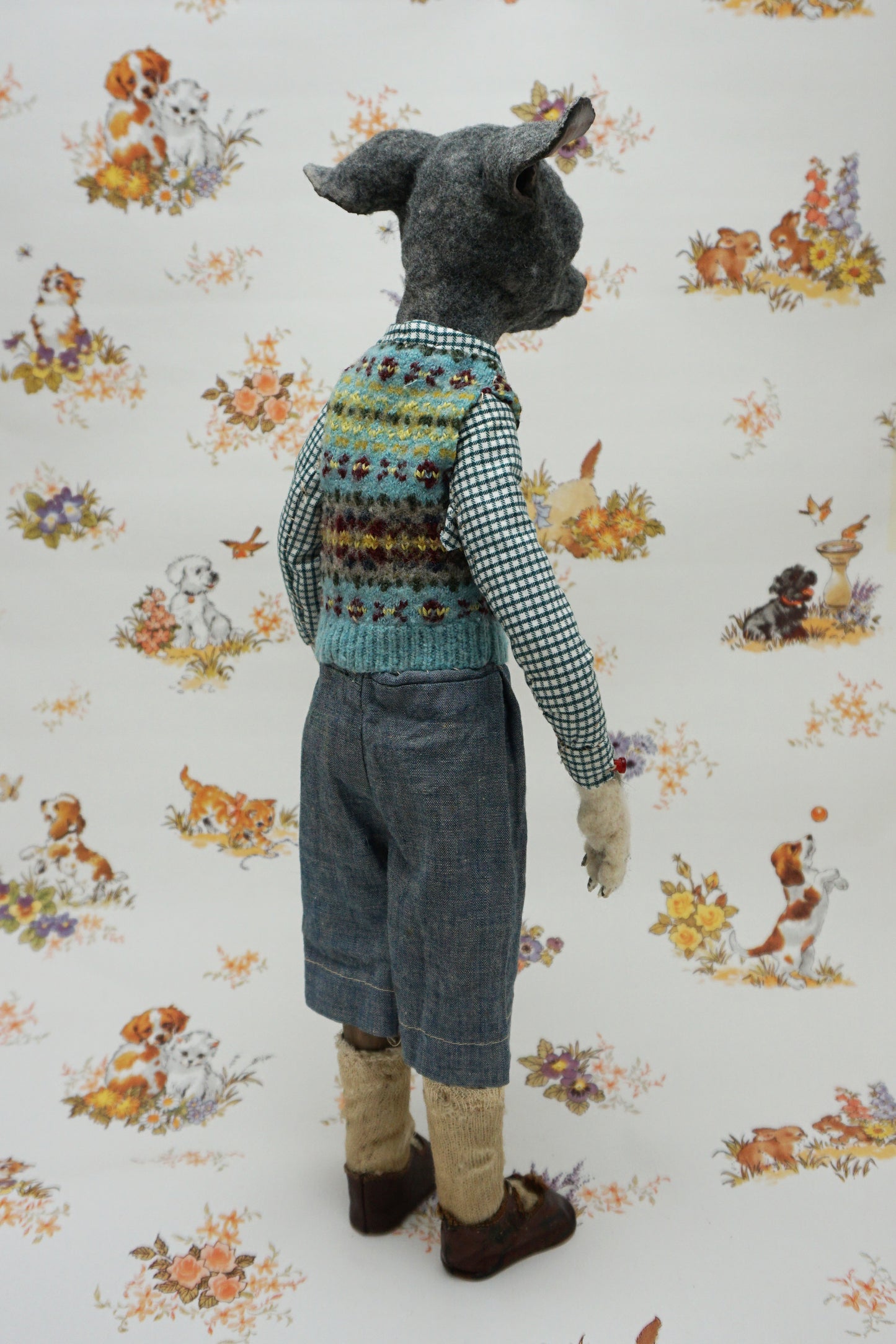 A whimsical, anthropomorphic dog figure stands upright, dressed in a checked shirt, patterned vest, and denim shorts. The background features a vintage-style wallpaper with illustrations of playful dogs and floral motifs.