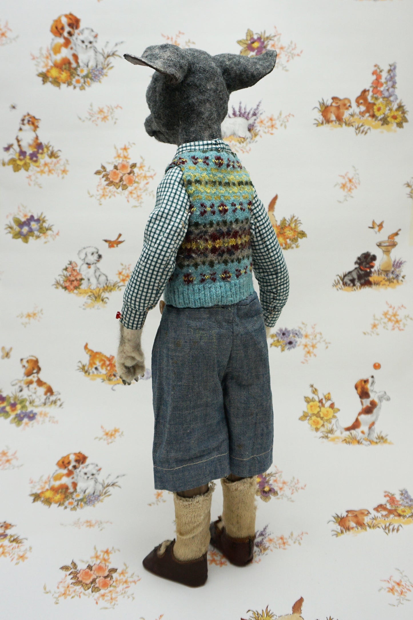 A whimsical, anthropomorphic dog figure stands upright, dressed in a checked shirt, patterned vest, and denim shorts. The background features a vintage-style wallpaper with illustrations of playful dogs and floral motifs.
