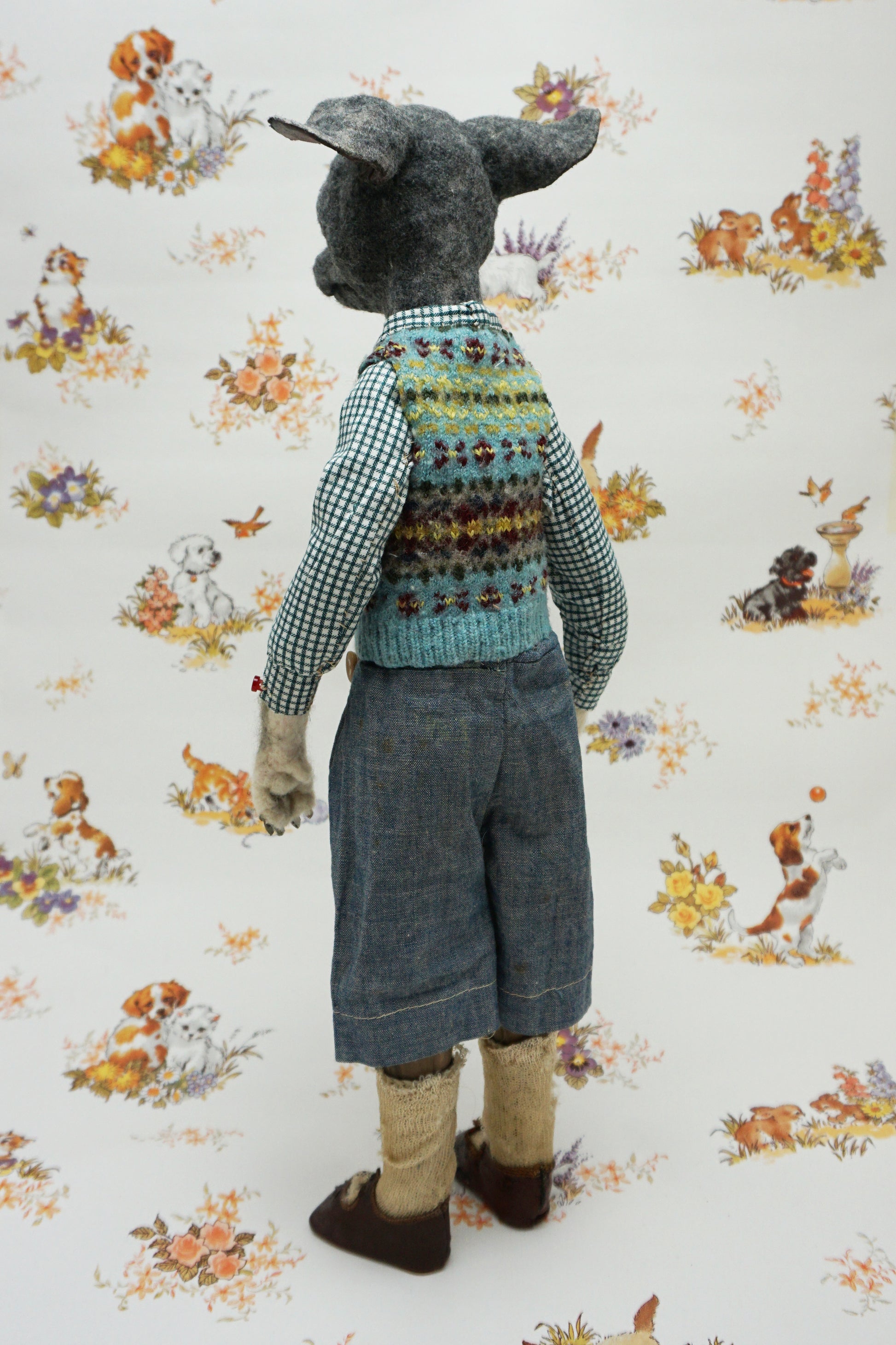 A whimsical, anthropomorphic dog figure stands upright, dressed in a checked shirt, patterned vest, and denim shorts. The background features a vintage-style wallpaper with illustrations of playful dogs and floral motifs.