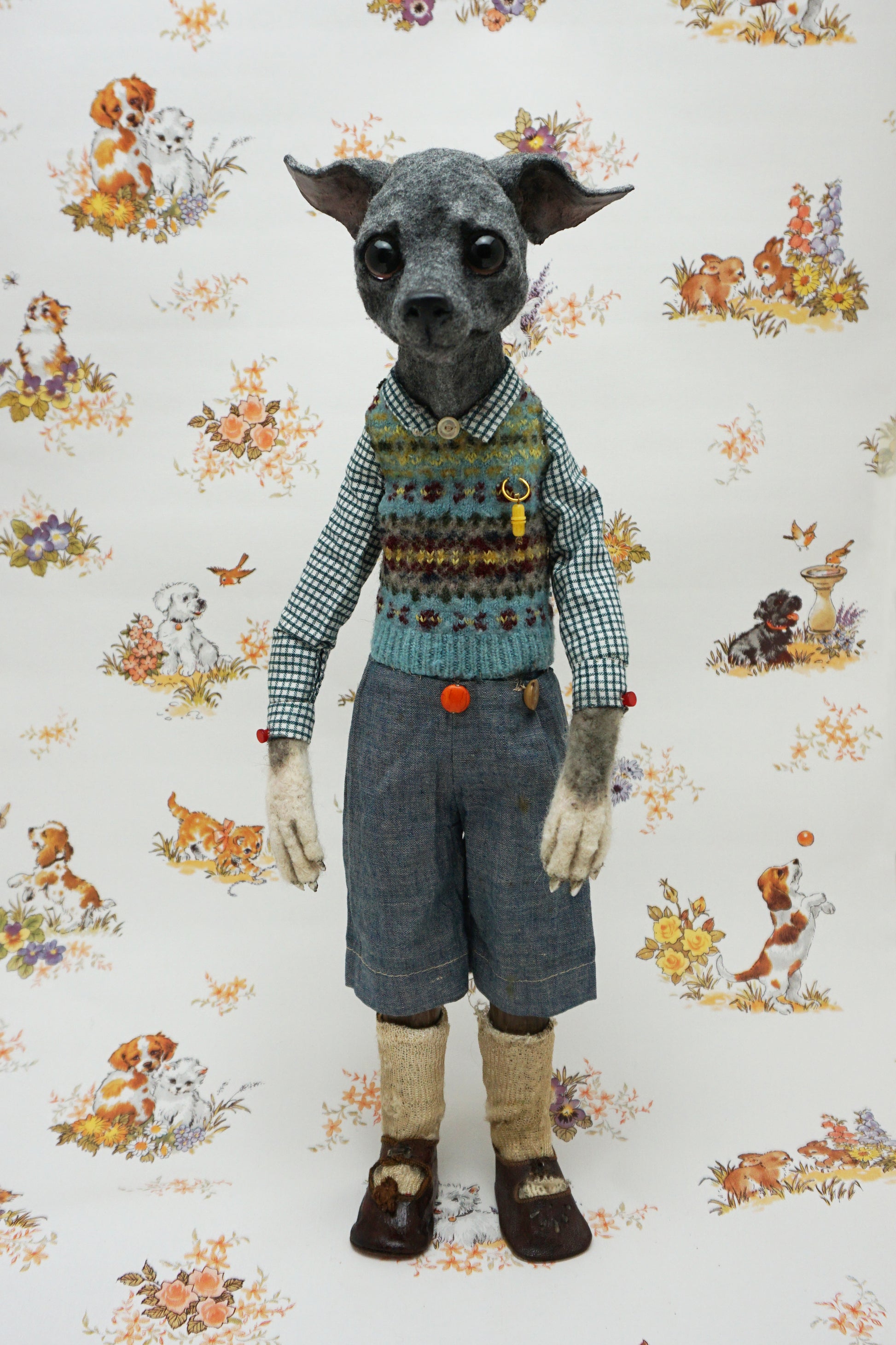 A whimsical, anthropomorphic dog figure stands upright, dressed in a checked shirt, patterned vest, and denim shorts. The background features a vintage-style wallpaper with illustrations of playful dogs and floral motifs.