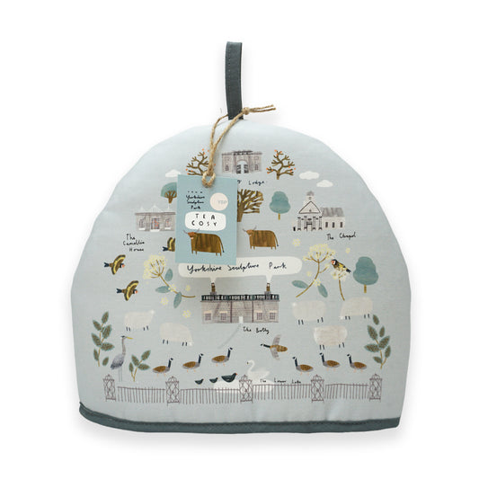 A tea cosy featuring a whimsical illustration of Yorkshire landscapes, including farms, trees, and animals like sheep and birds. The design is in soft pastel colours, with a grey trim and a tag attached, adding a charming touch to any kitchen.