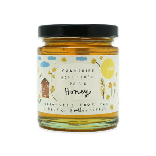 A glass jar of Yorkshire Sculpture Park Honey with a black lid and a cream-coloured label featuring yellow flowers, a bee hive, and blue clouds. The honey inside is golden amber, and a yellow circular badge on the label reads 'YSP'.