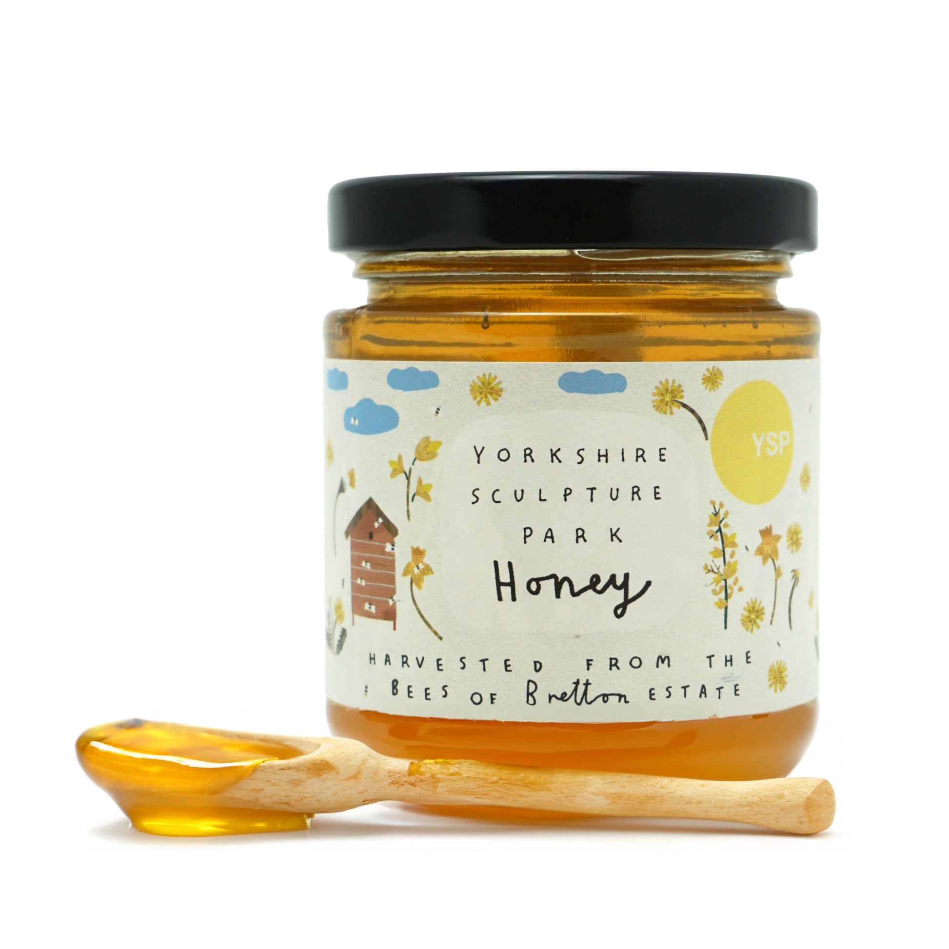 A jar of Yorkshire Sculpture Park honey with a black lid and a decorative label featuring clouds and flowers. A wooden honey dipper rests beside the jar, showcasing the golden honey inside. The label notes it is harvested from the Bees of Bretton Estate.